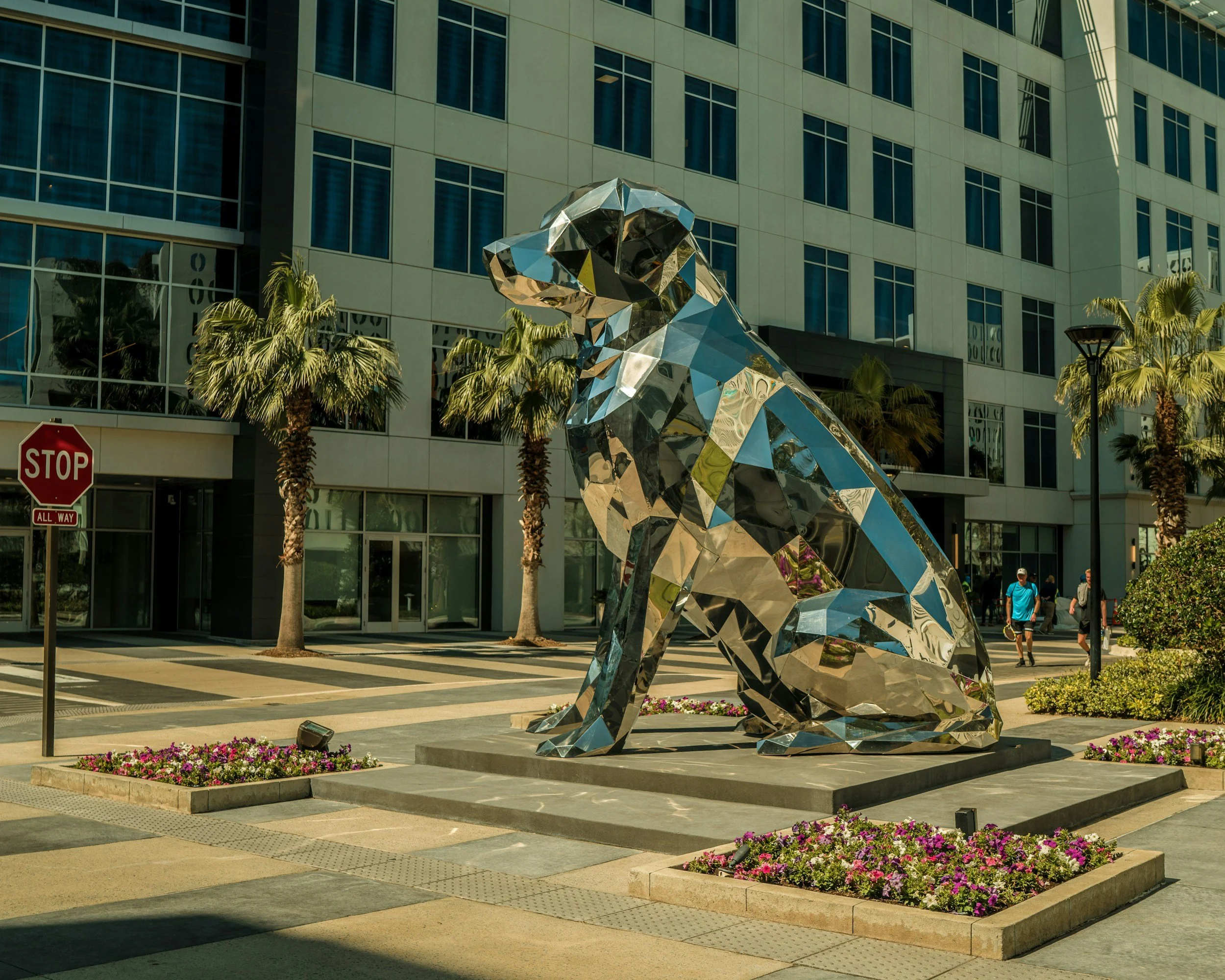 A metallic sculpture of a sitting dog in front of a modern glass building. The sculpture resembles a dog with an abstract, geometric design. There are palm trees and pink flowers surrounding the sculpture, and a stop sign visible on the street corner.