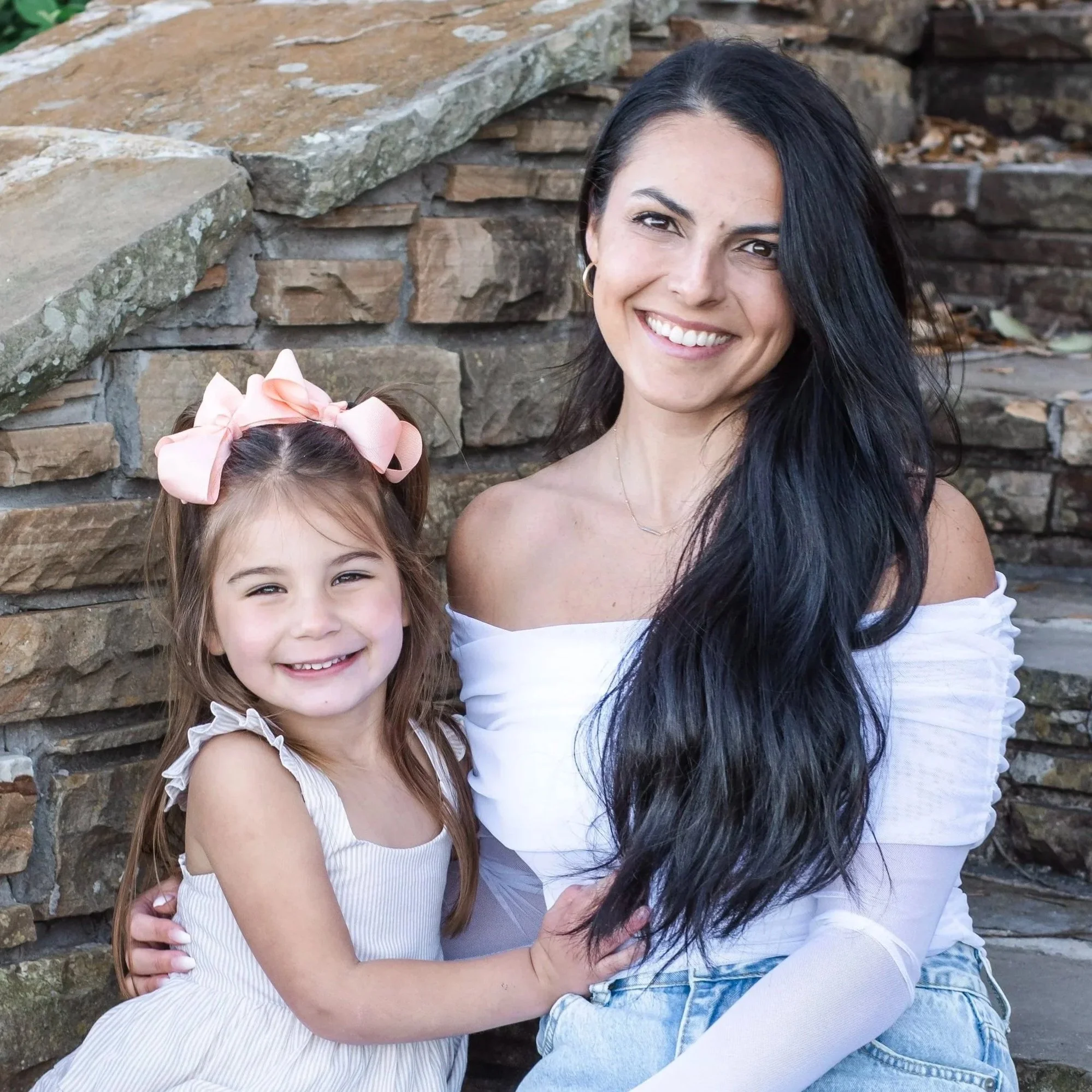 A woman with long black hair and hoop earrings smiling, and a young girl with light brown hair and a pink bow, sitting together outdoors in front of a stone wall.