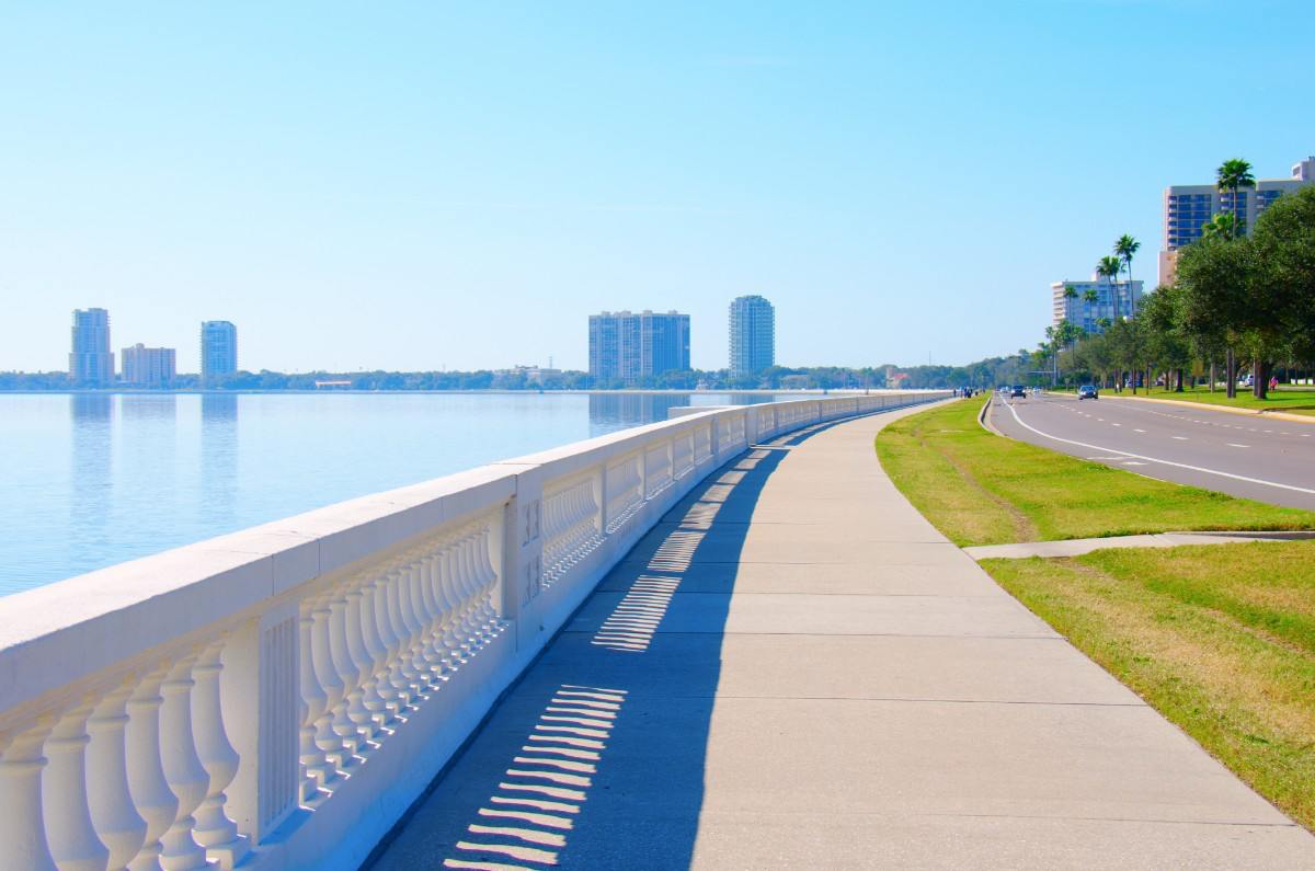 A waterfront promenade with a white railing on the left, a grassy strip with trees, and a road with cars on the right, with a city skyline of tall buildings across the water in the background.
