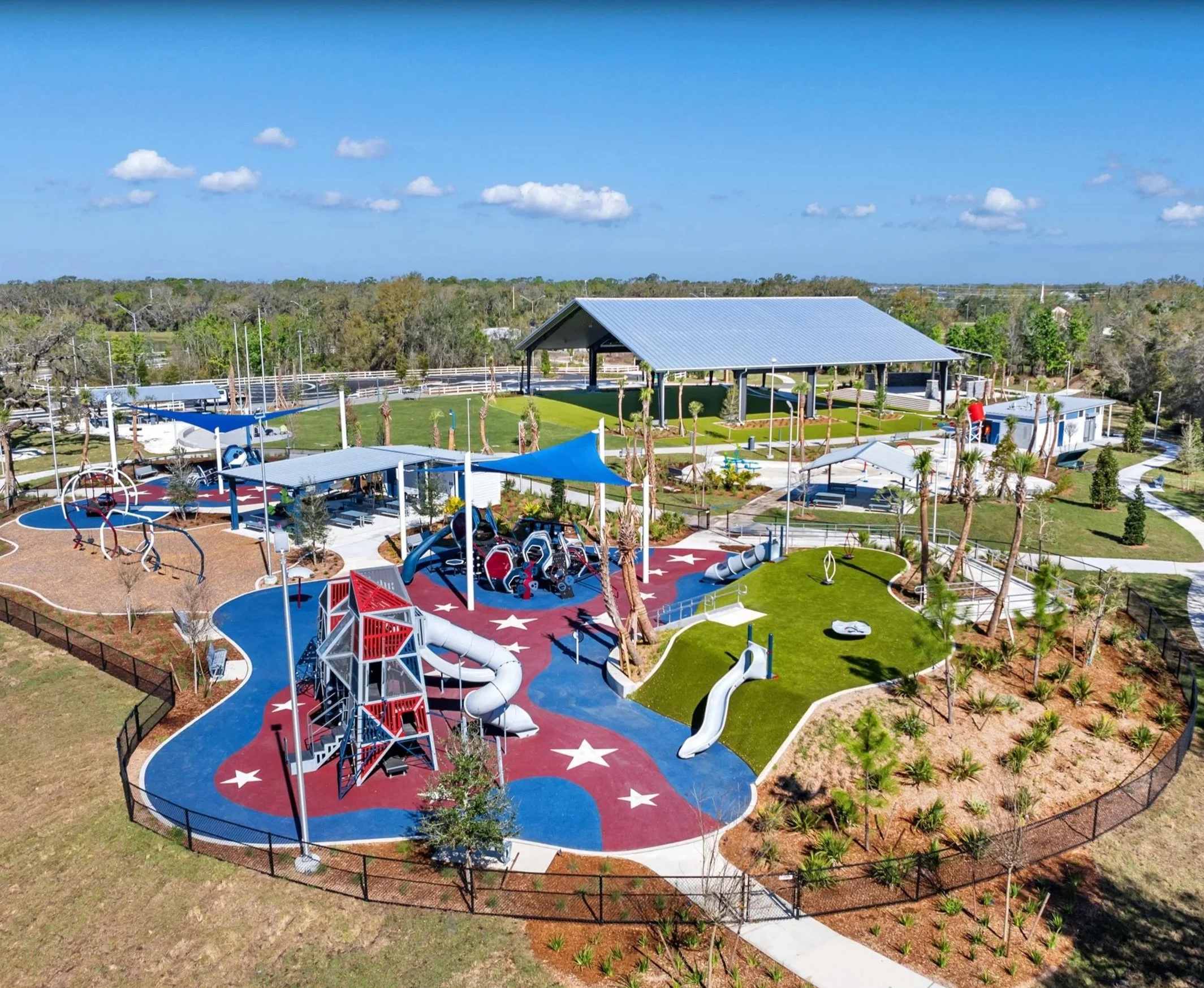An aerial view of a colorful outdoor playground with slides, climbing structures, and a splash pad, surrounded by green grass and trees under a partly cloudy sky.