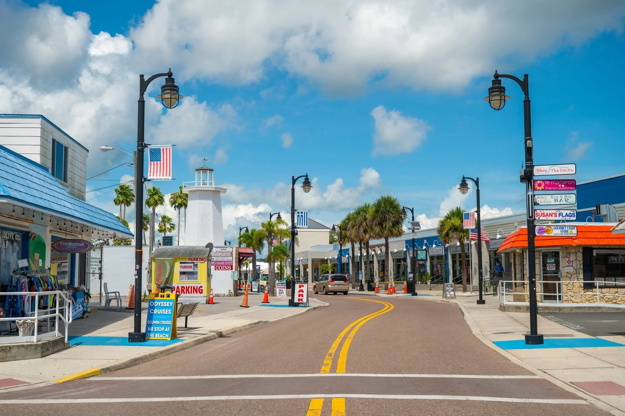 A street scene in a shopping area with palm trees, signs, a lighthouse, and storefronts under a partly cloudy sky.