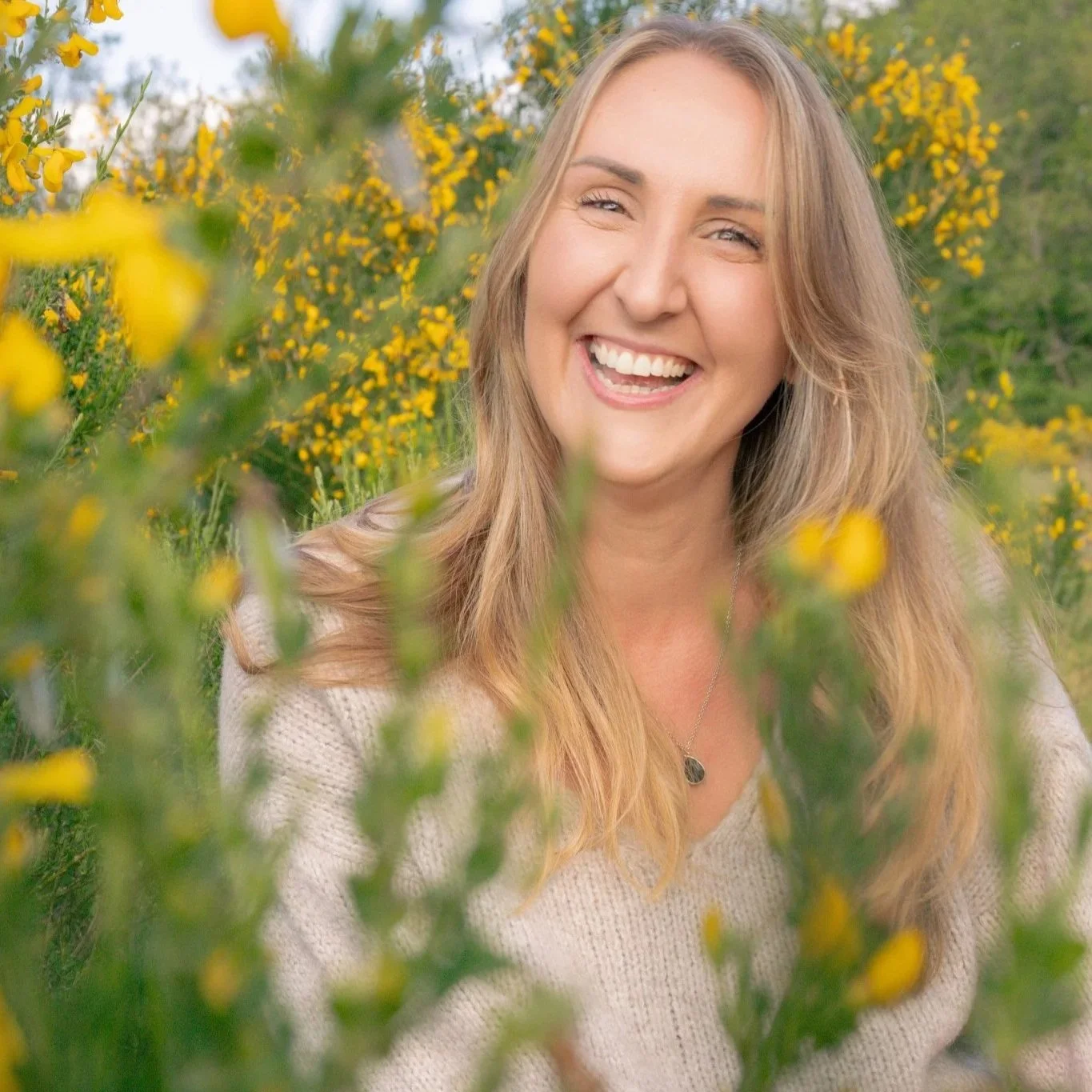 A smiling woman with long, wavy blonde hair in a field of yellow flowers, wearing a light-colored sweater and a necklace, with green trees and a cloudy sky in the background.