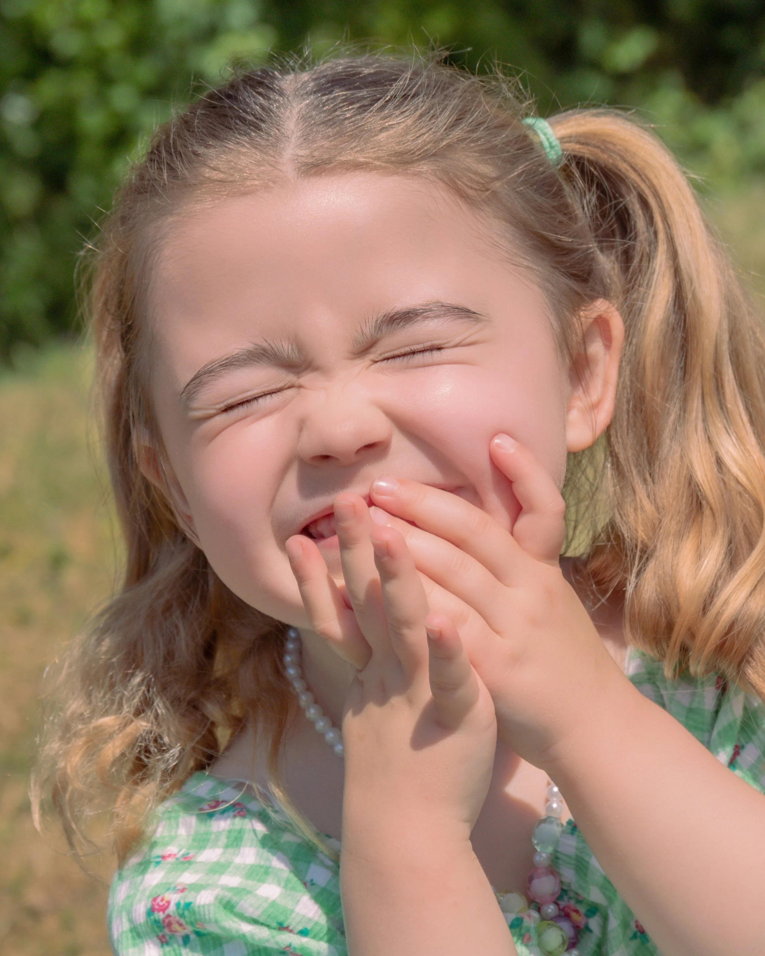 A young girl with curly, light brown hair in pigtails, smiling and covering her mouth with her hand, outdoors with greenery in the background, wearing a green checkered dress and pearl jewelry.