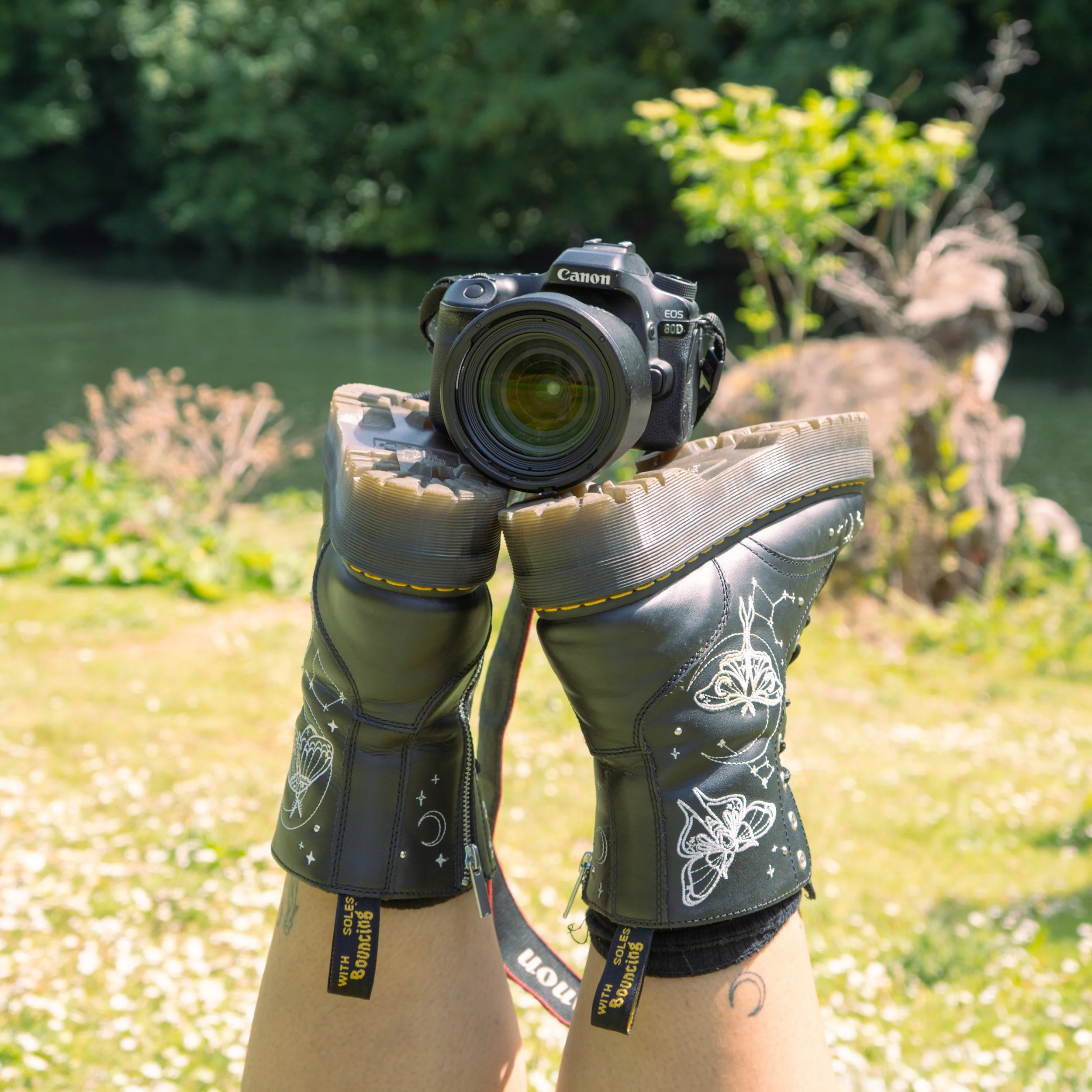 Close-up of shoes  with black jackets with butterfly and flower designs, outdoors in a sunny, wooded area near a body of water, balancing a Canon camera.