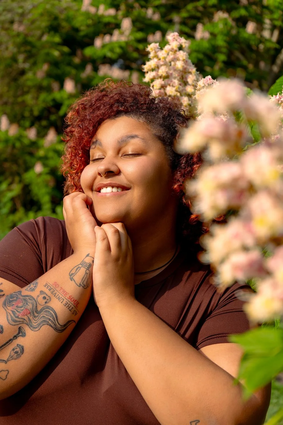A woman with curly red-brown hair and tattoos, smiling with her eyes closed, in a garden with blooming pink and white flowers.