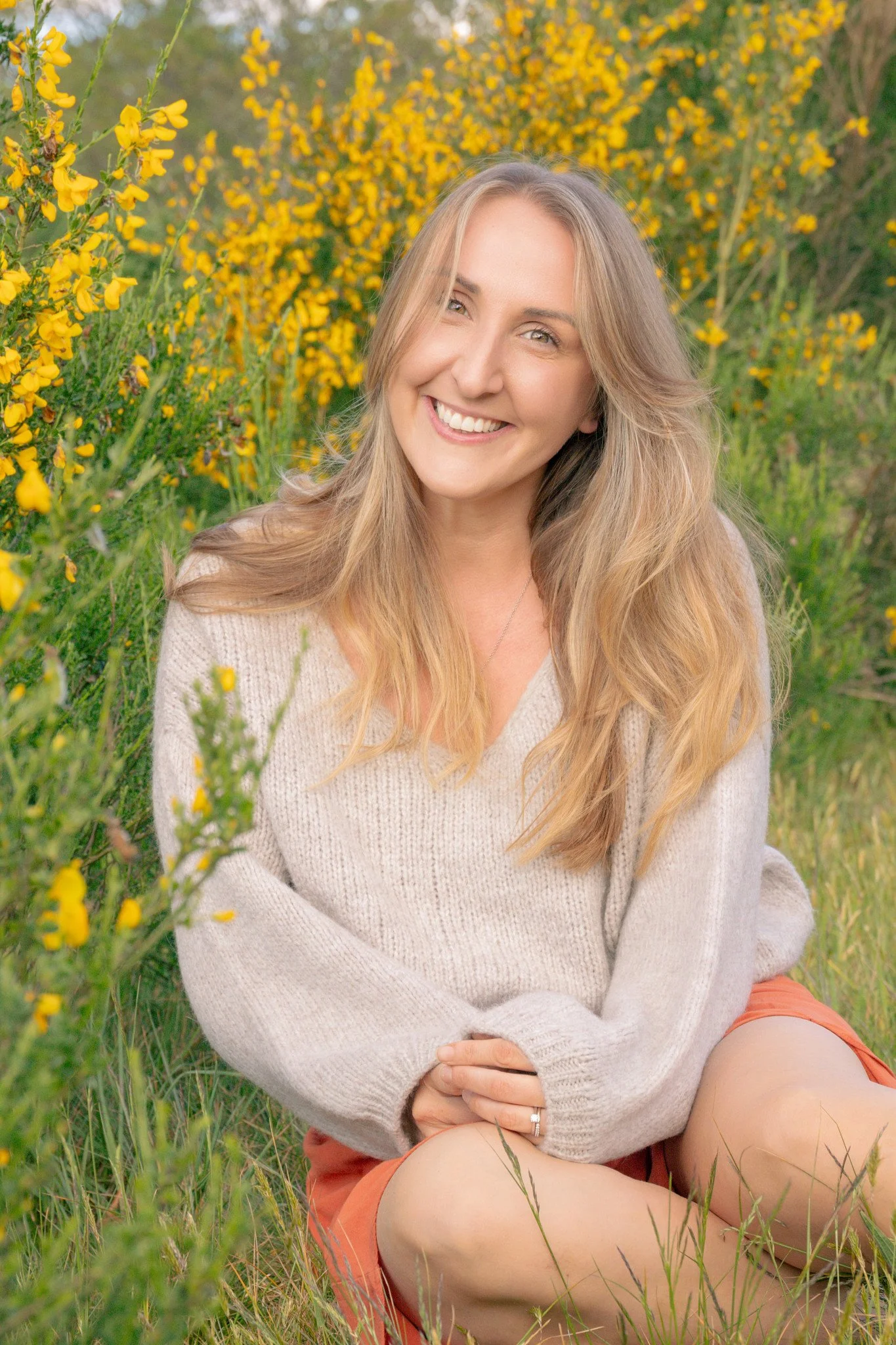 A smiling woman sitting on the grass among yellow wildflowers outdoors.