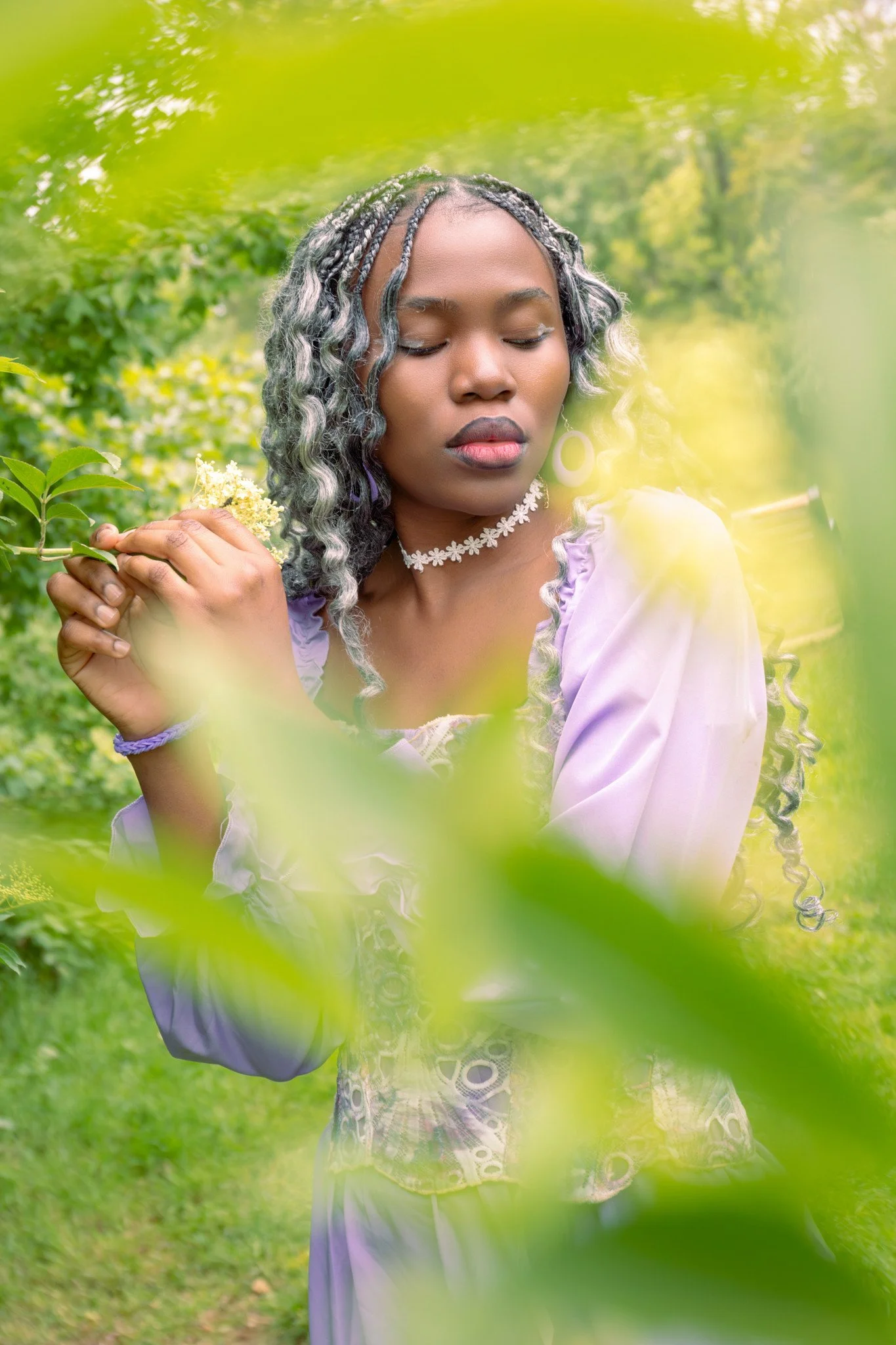 A woman with curly, gray-tinted hair and dark skin, wearing a lavender top, floral choker, and large hoop earrings, standing outdoors among greenery with her eyes closed and a peaceful expression.