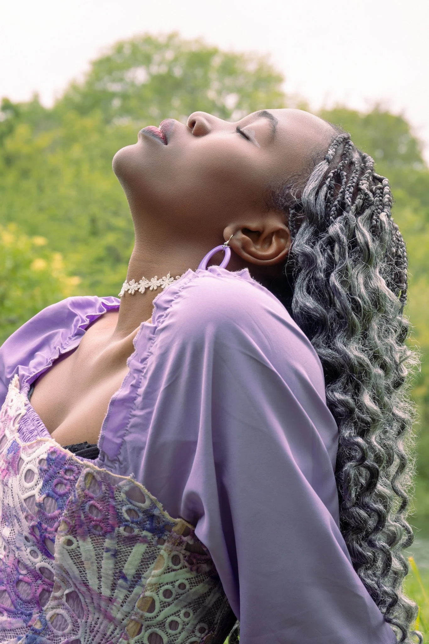 Side view of a woman with closed eyes, braids with silver highlights, wearing a purple ruffled top with lace details, outdoors with green trees in the background.