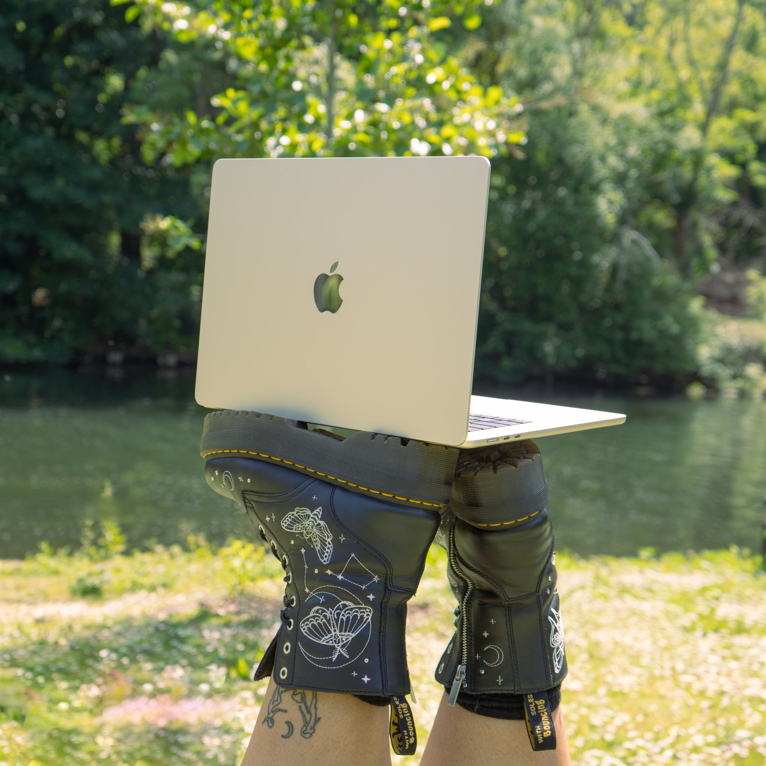 Close-up of a person wearing black pants with white shell patterns and a black jacket with celestial designs, standing outdoors with a white Apple computer on their shoulder and greenery in the background.
