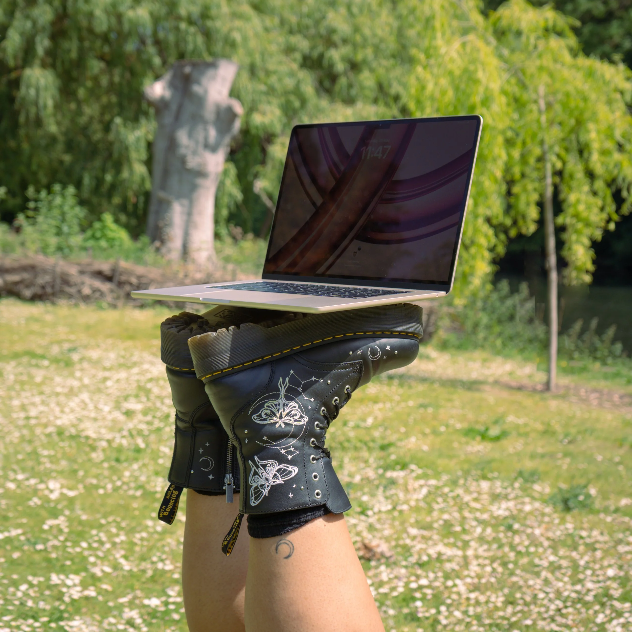 Close-up of a person's leg wearing a black leather glove with patches of white floral embroidery, holding a silver laptop outdoors with green trees and white flower petals in the background.