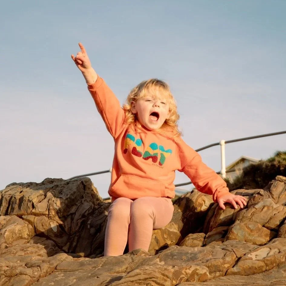 A young girl with curly blonde hair sitting on rocks outdoors, wearing an orange sweatshirt and pink pants, shouting with her right arm raised and finger pointing upwards.