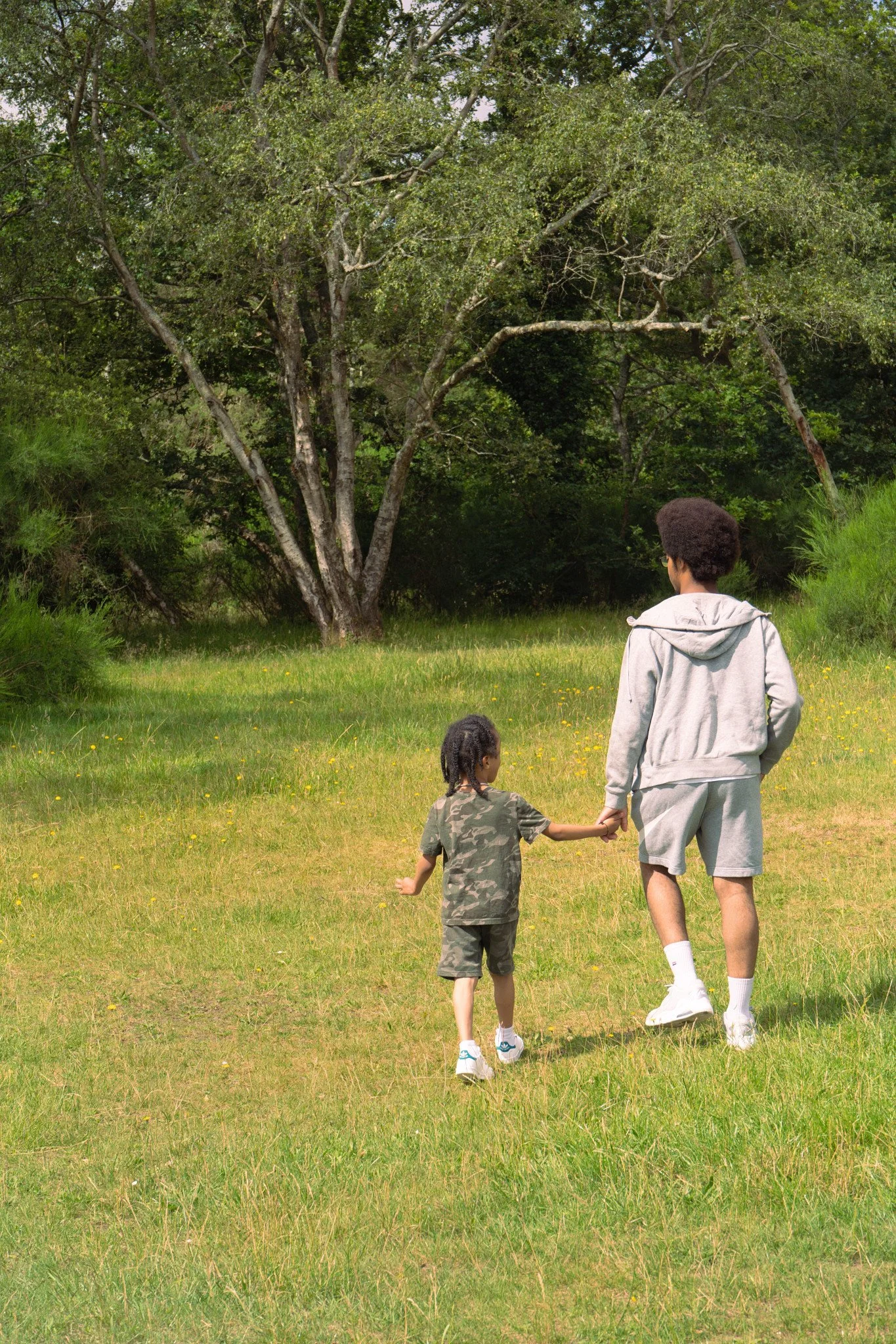 hild holding an older sibling’s hand while walking through a sunny meadow, family photography session in Burnham Beeches