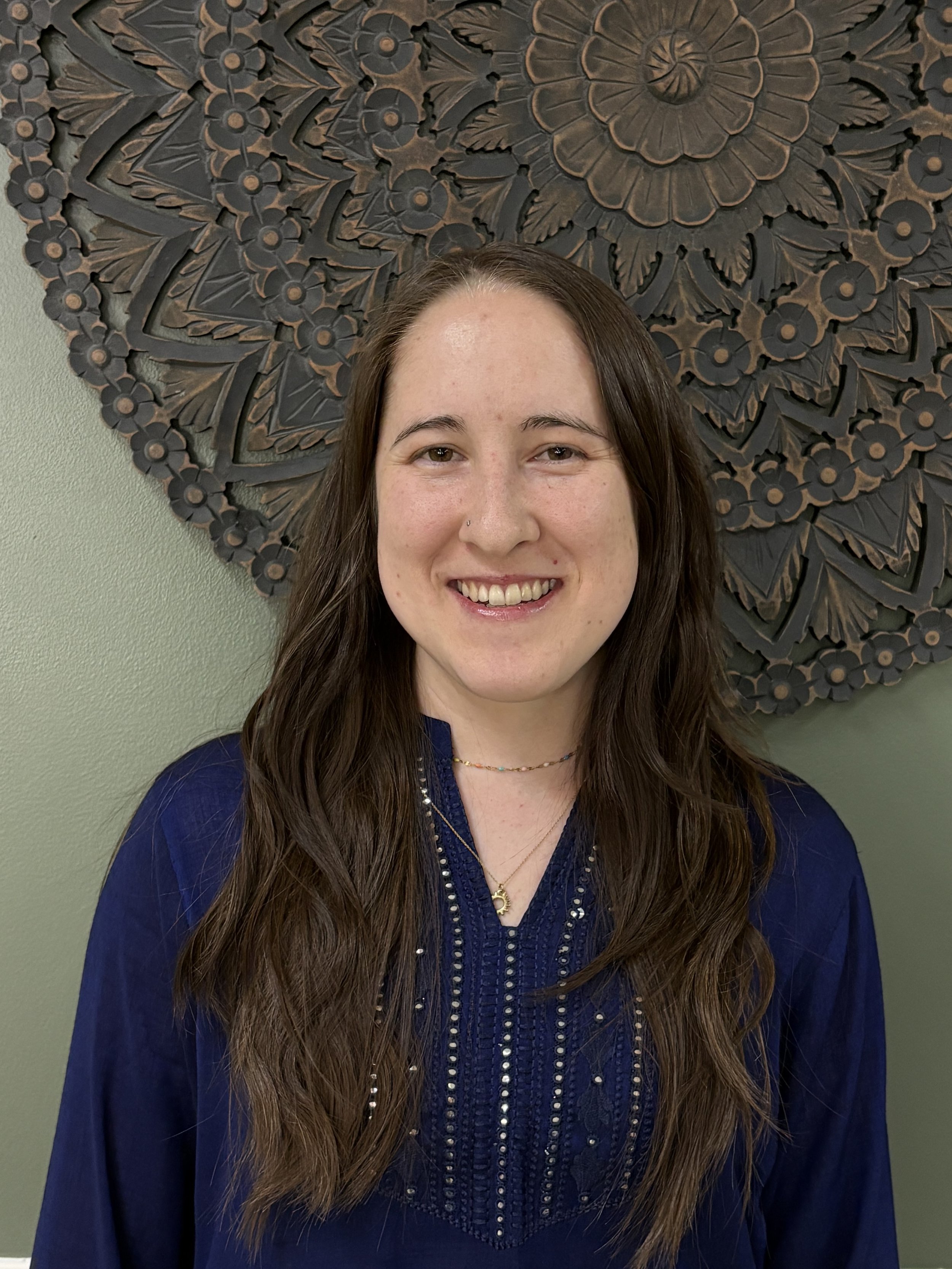 A woman with long brown hair, smiling, standing in front of a large decorative wooden wall art piece with floral patterns.