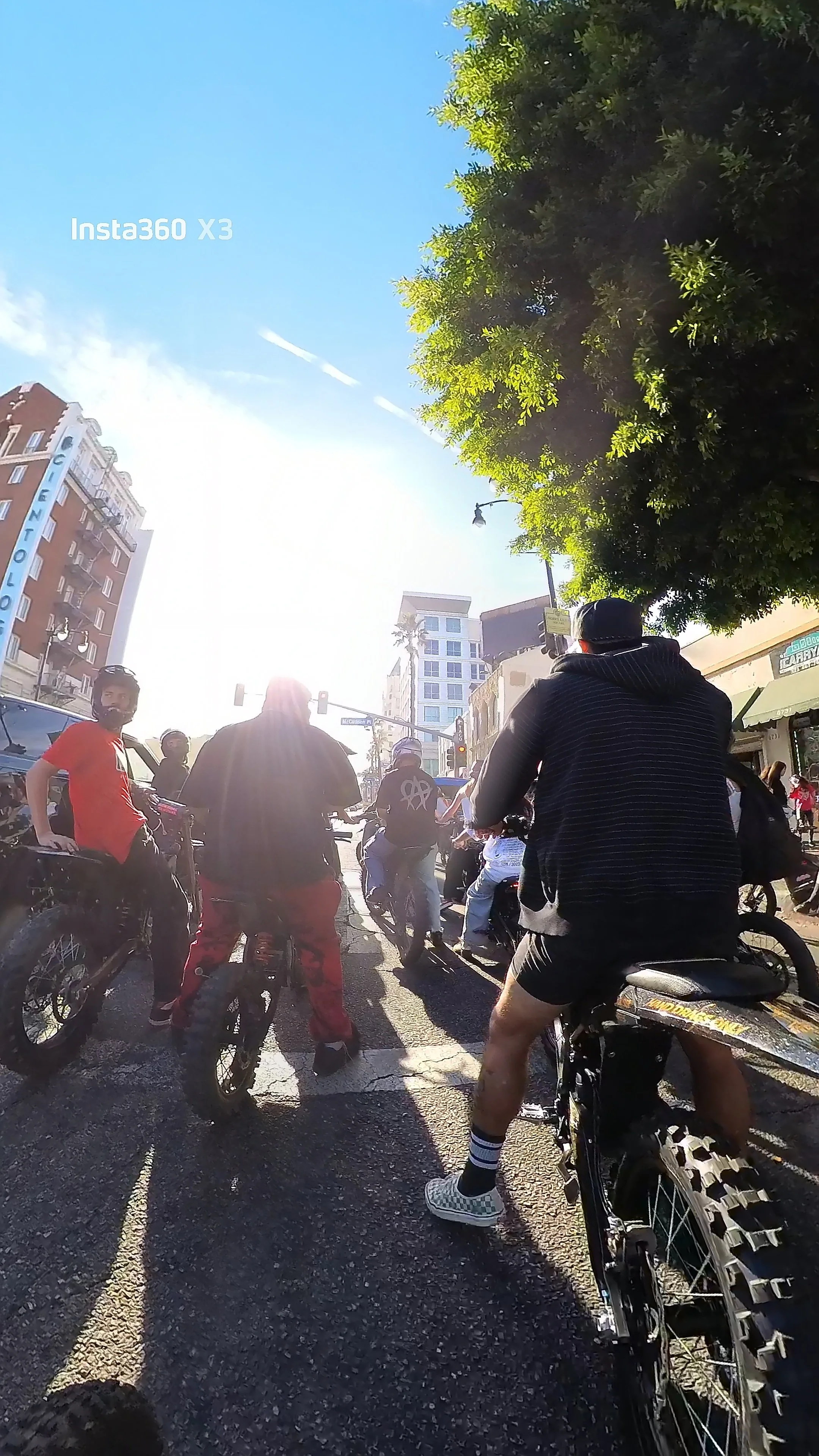 Group of people riding bikes on a city street on a sunny day, with tall buildings and trees visible in the background.