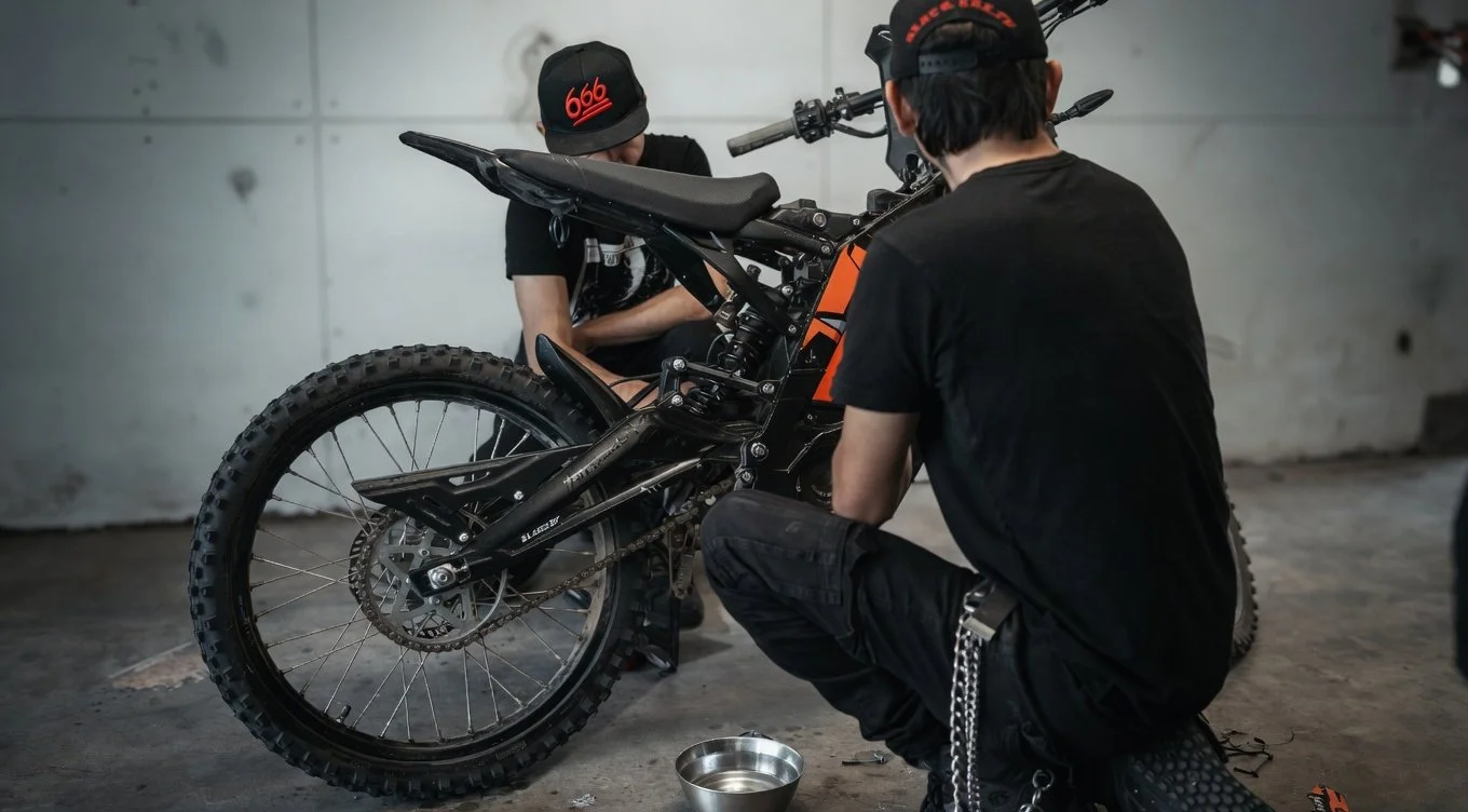 Two individuals working on a dirt bike in an indoor setting. One person is kneeling while the other is standing, both focused on the bike.