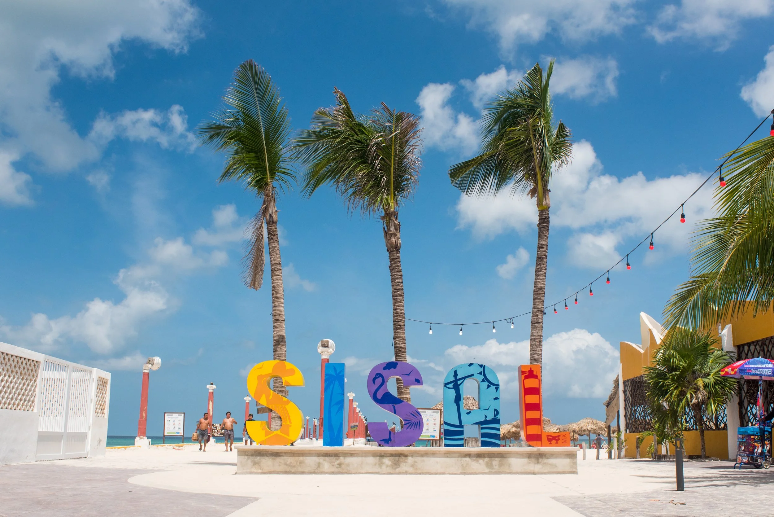Colorful large letters spelling "SISAL" beach with palm trees, string lights, and a blue sky with clouds. Yucatán, México The Green Maya Project