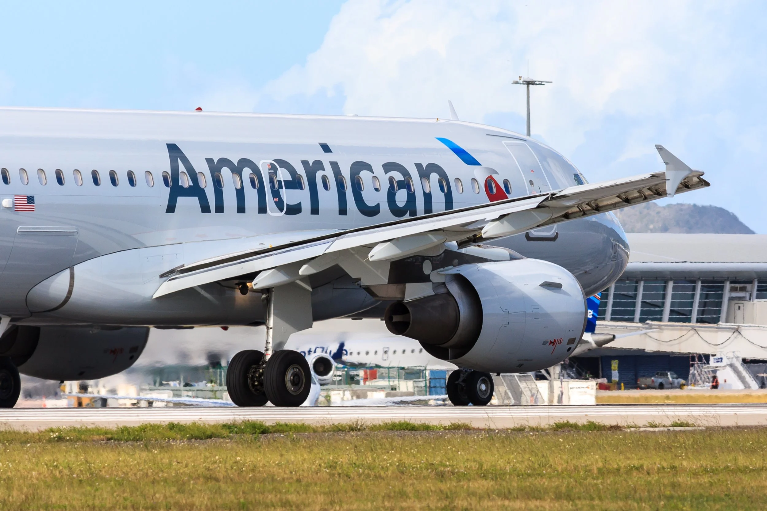 Close-up of an American Airlines airplane on the tarmac at an airport, showing part of the fuselage, landing gear, and engine with a background of cloudy sky and airport structures.