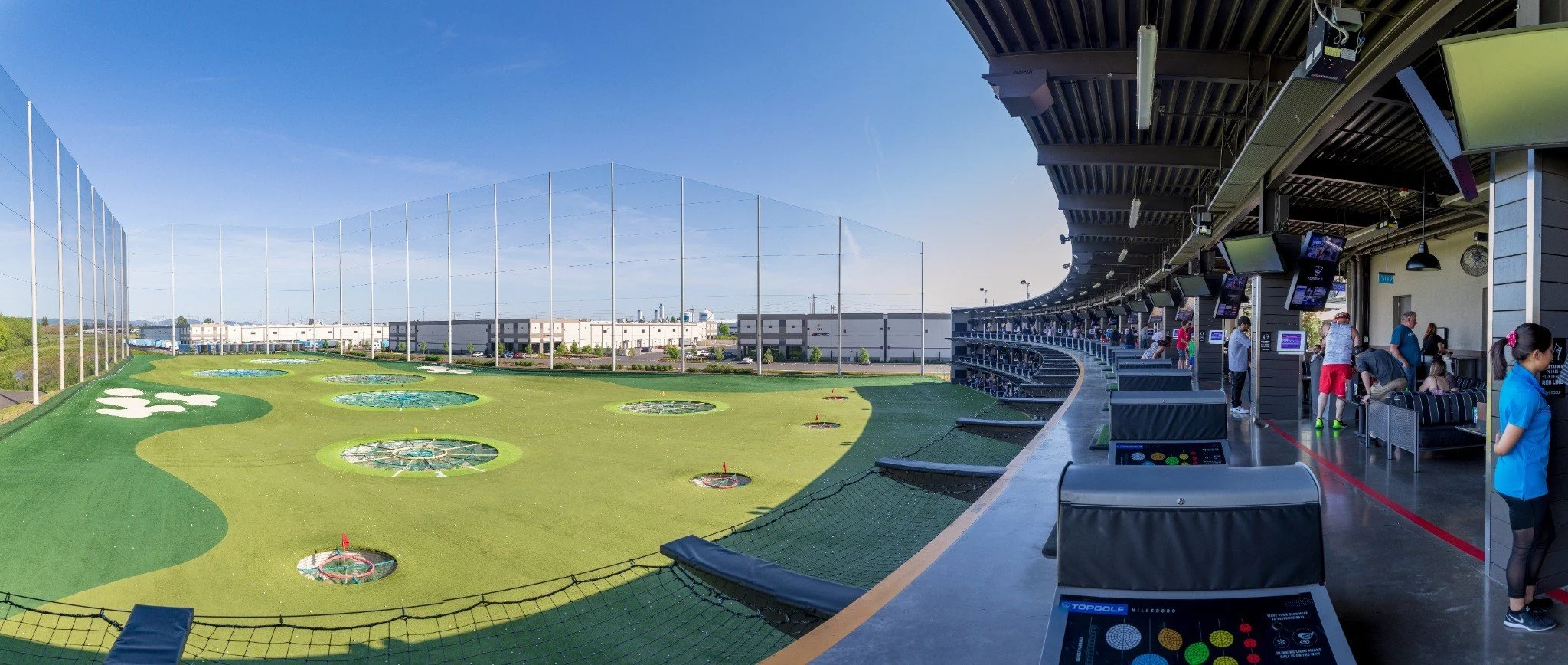 Indoor Topgolf golf driving range with people playing and watching, computer screens, and a large open outdoor field with multiple batting cages.