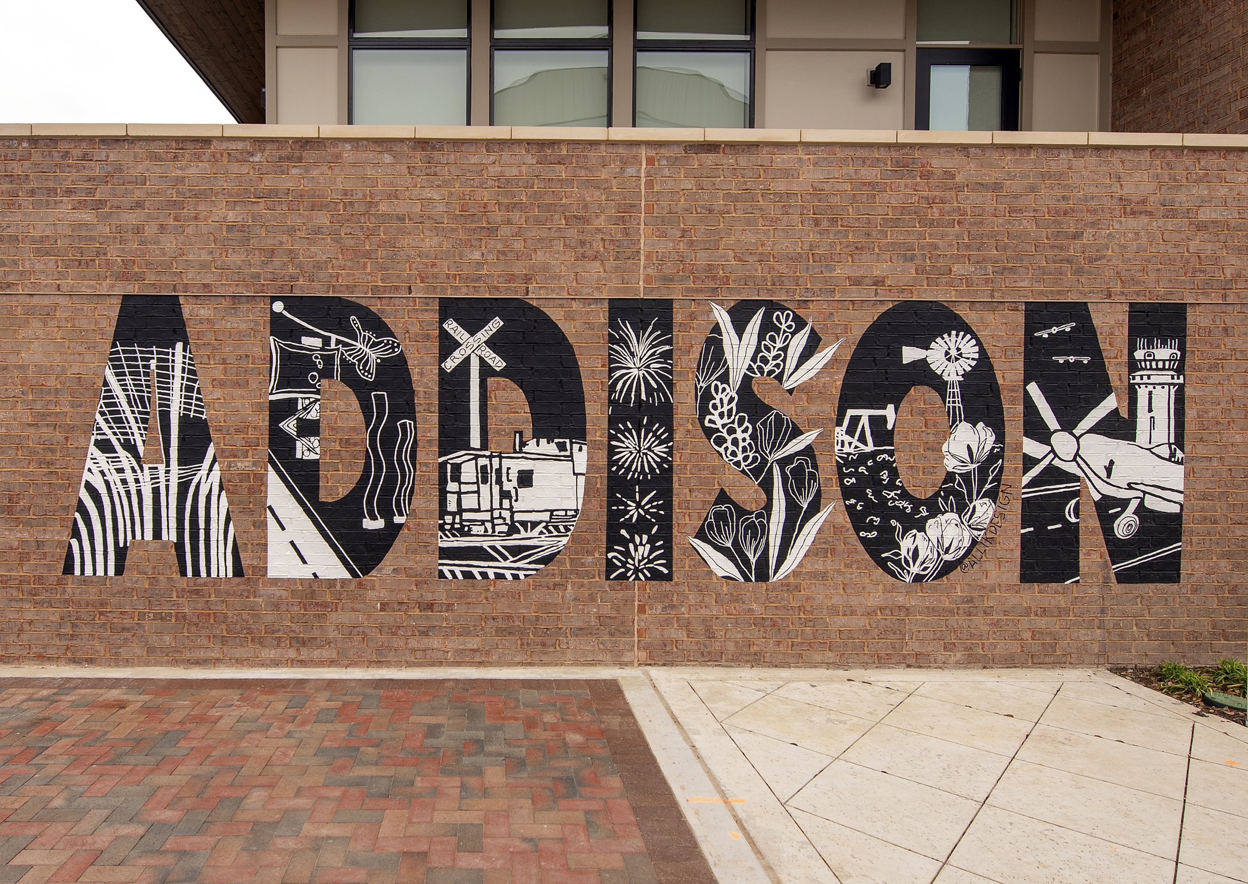 A brick wall with black and white murals of various objects and scenes, including trees, a butterfly, fireworks, and a tower.