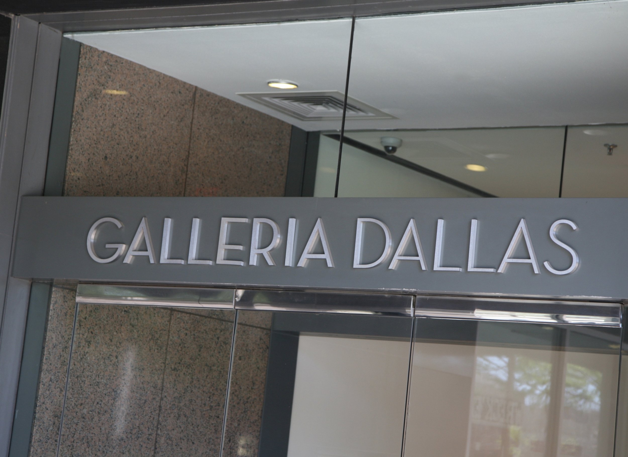 Part of a sign reading 'GALLERIA DALLAS' in metallic letters, mounted on a wall inside a building with tile and glass surfaces.