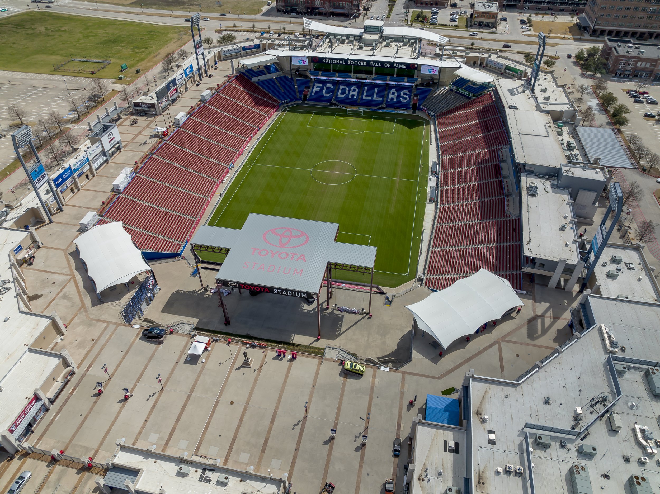 Aerial view of Toyota Stadium, home of FC Dallas, showing the soccer field with red empty seats in the stands, and surrounding parking lots and urban buildings.