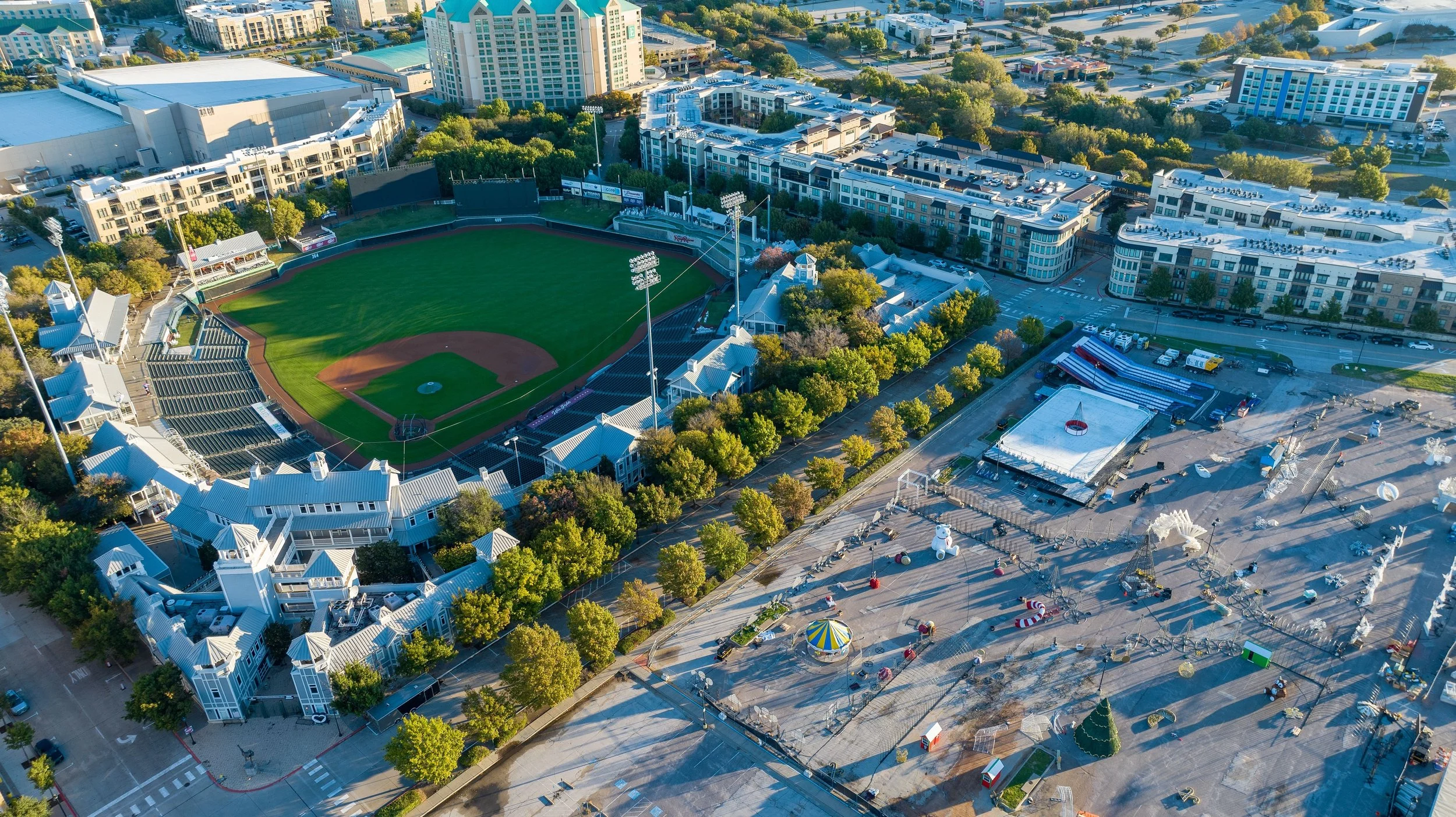 An aerial view of a cityscape with a baseball stadium at the center, surrounded by residential and commercial buildings, trees, and an amusement park area with rides and attractions.