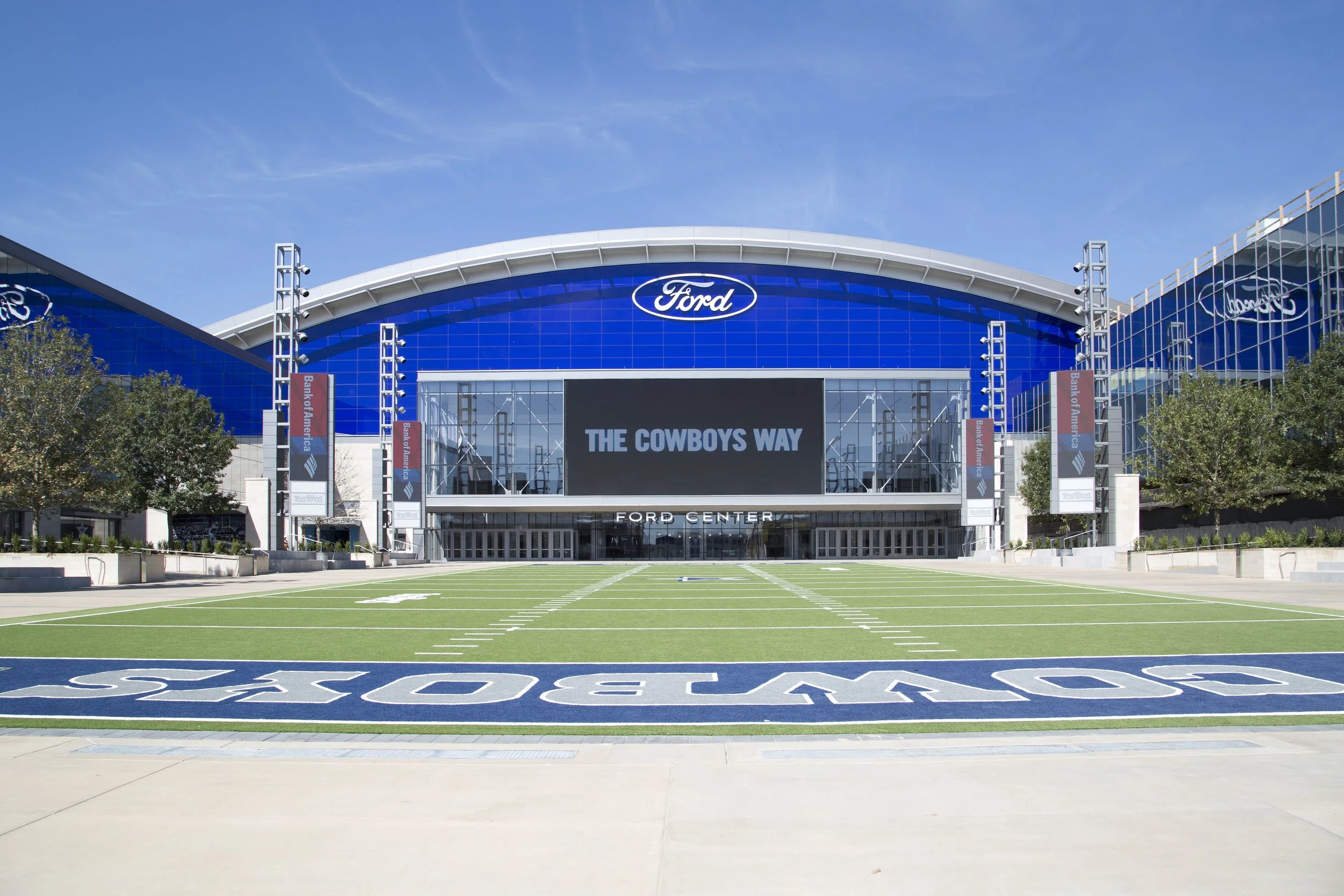 Exterior view of Ford Center with a football field in front, large digital screen displaying 'THE COWBOYS WAY', and blue building with Ford logos.