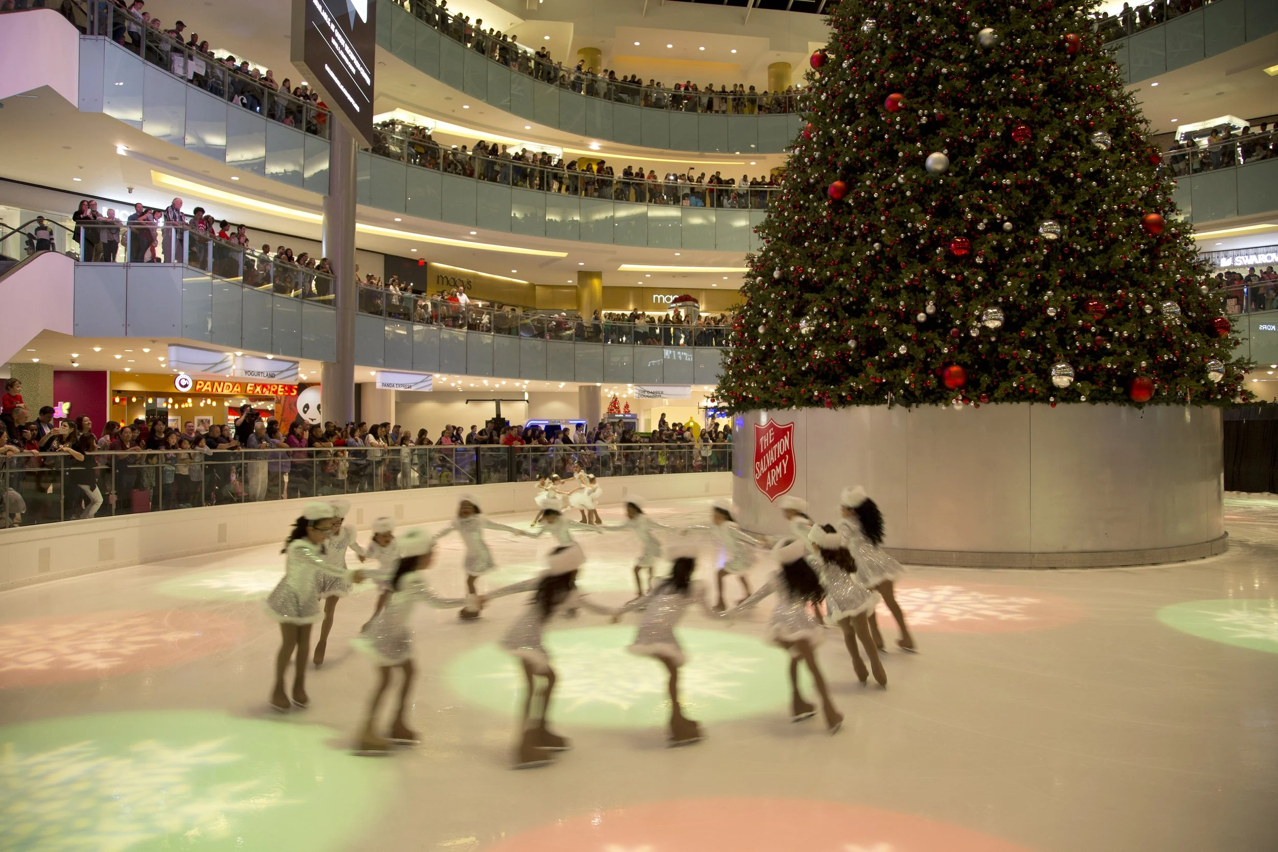 Indoor ice skating rink at a shopping mall decorated for Christmas with a large Christmas tree and skaters on the ice. Multiple levels of the mall are visible with people watching from the balconies.
