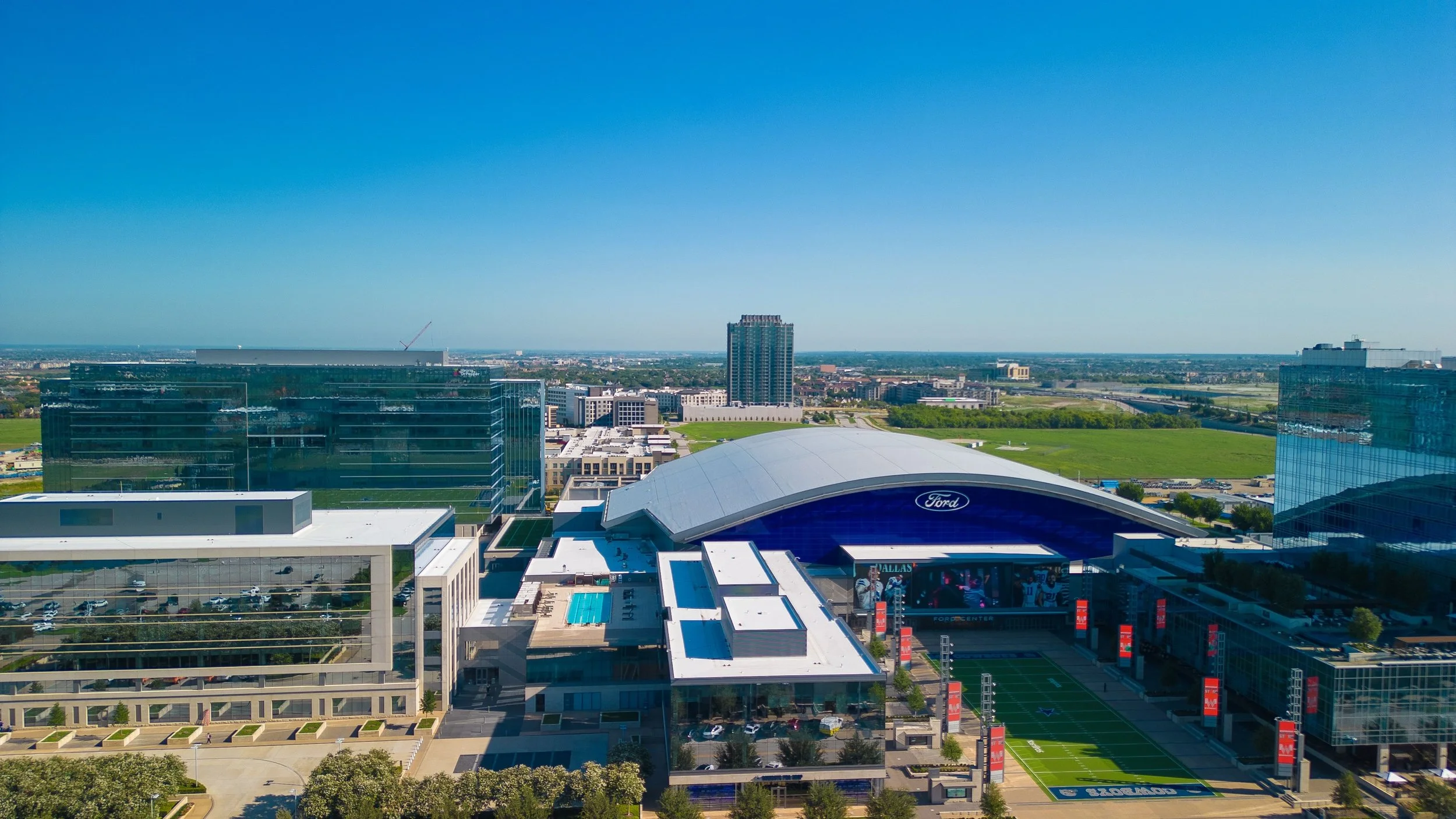 Aerial view of a cityscape featuring modern glass buildings, a large stadium with a Ford logo, a swimming pool, green fields, and a clear blue sky.