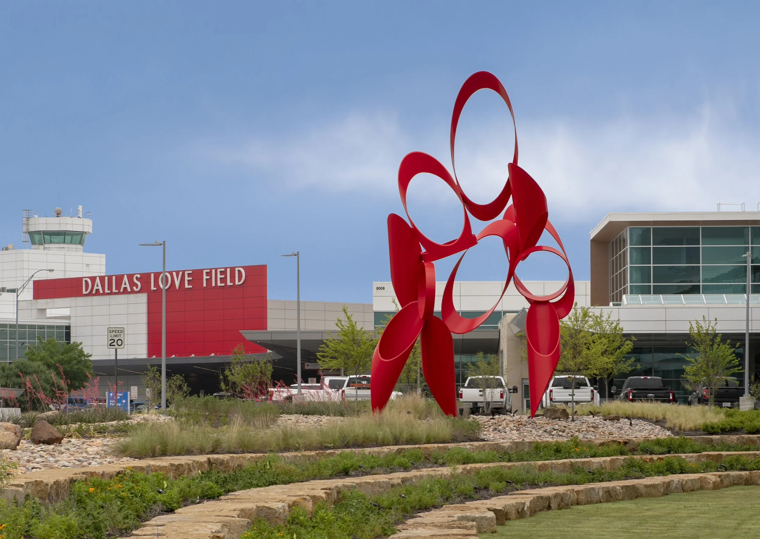 Large red abstract sculpture in front of Dallas Love Field airport terminal, with building and control tower visible in background, under a blue sky.