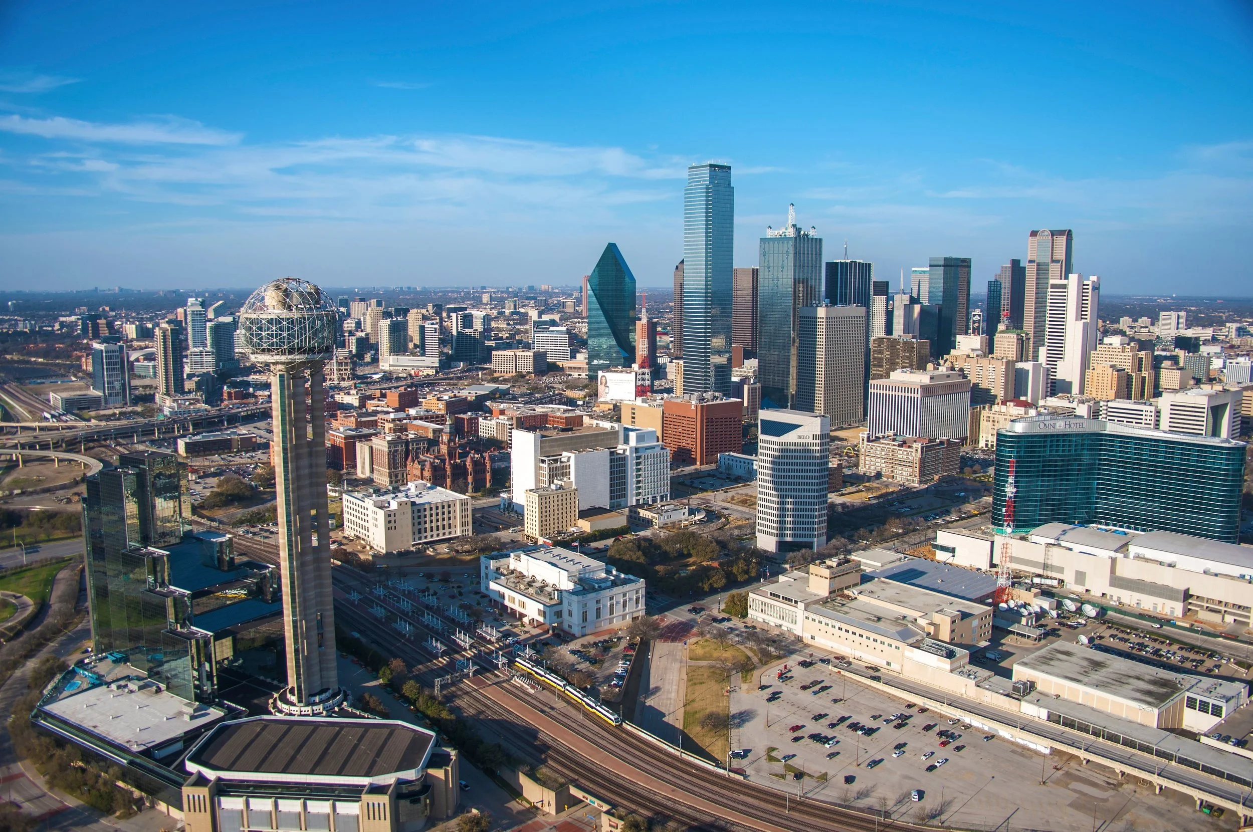 Aerial view of downtown Dallas, Texas, featuring skyscrapers, including the Bank of America Plaza and the Reunion Tower, under a blue sky with scattered clouds.