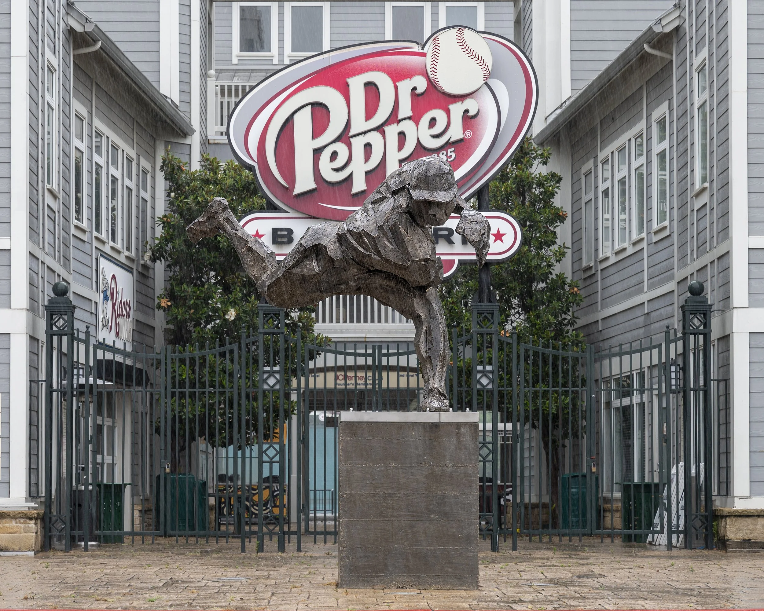 Image of a statue of a boy batting, in front of a gate with a Dr. Pepper sign, and houses or buildings in the background.