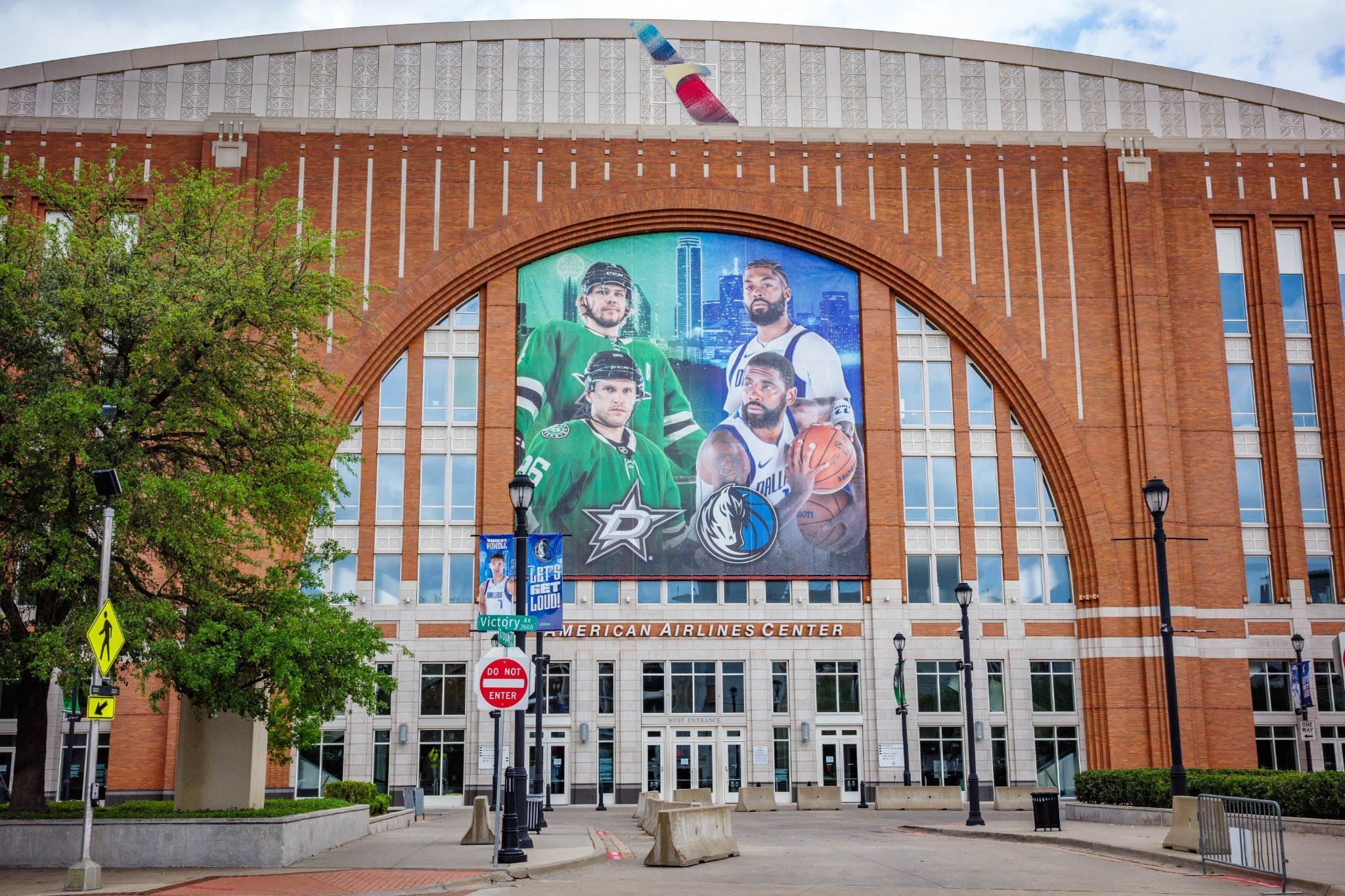 Exterior view of the American Airlines Center building with large poster of Dallas Stars hockey team players displayed on the facade.