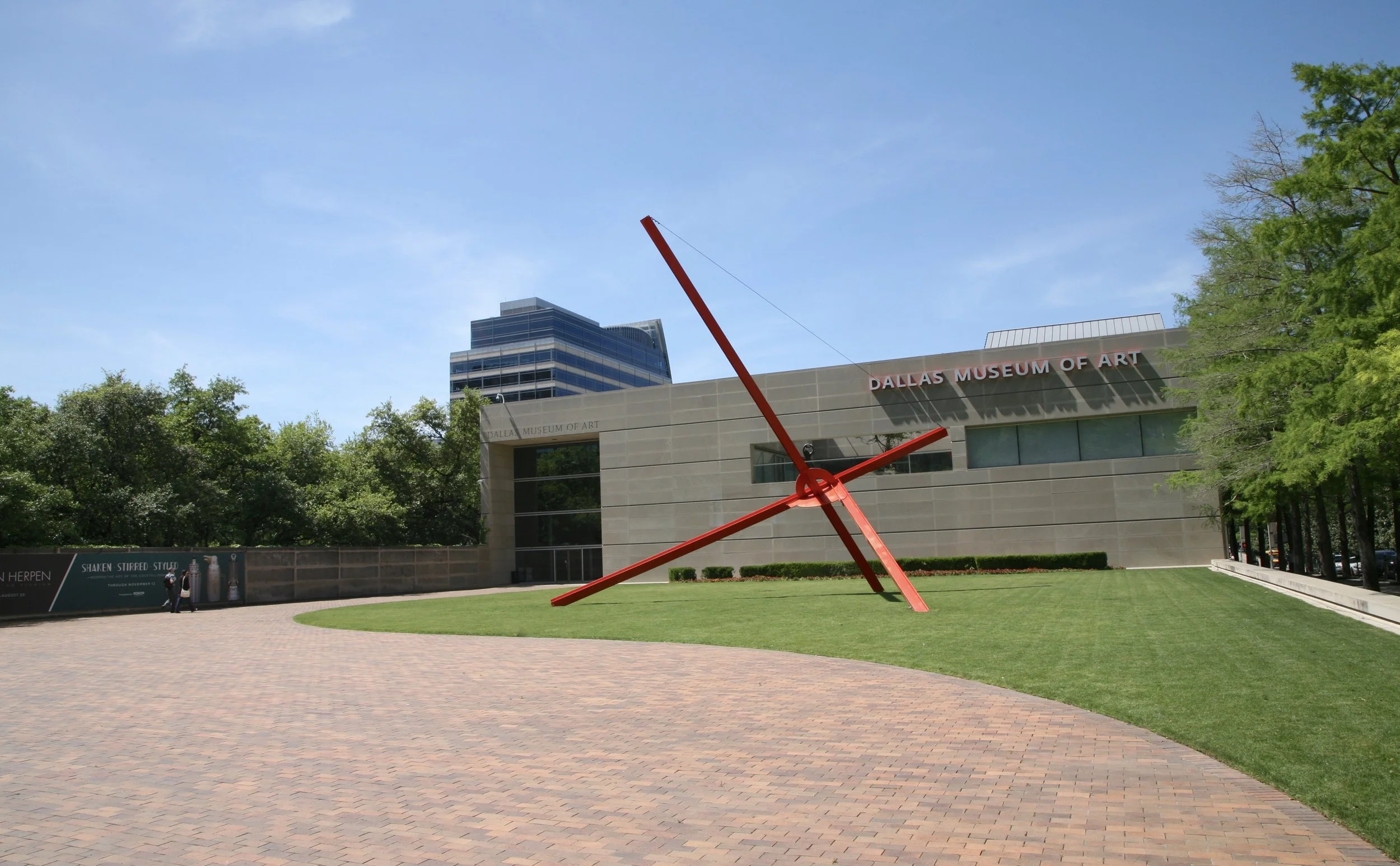 Exterior view of the Dallas Museum of Art with a large red outdoor clock sculpture in the foreground, trees lining the area, and a modern high-rise building behind the museum.