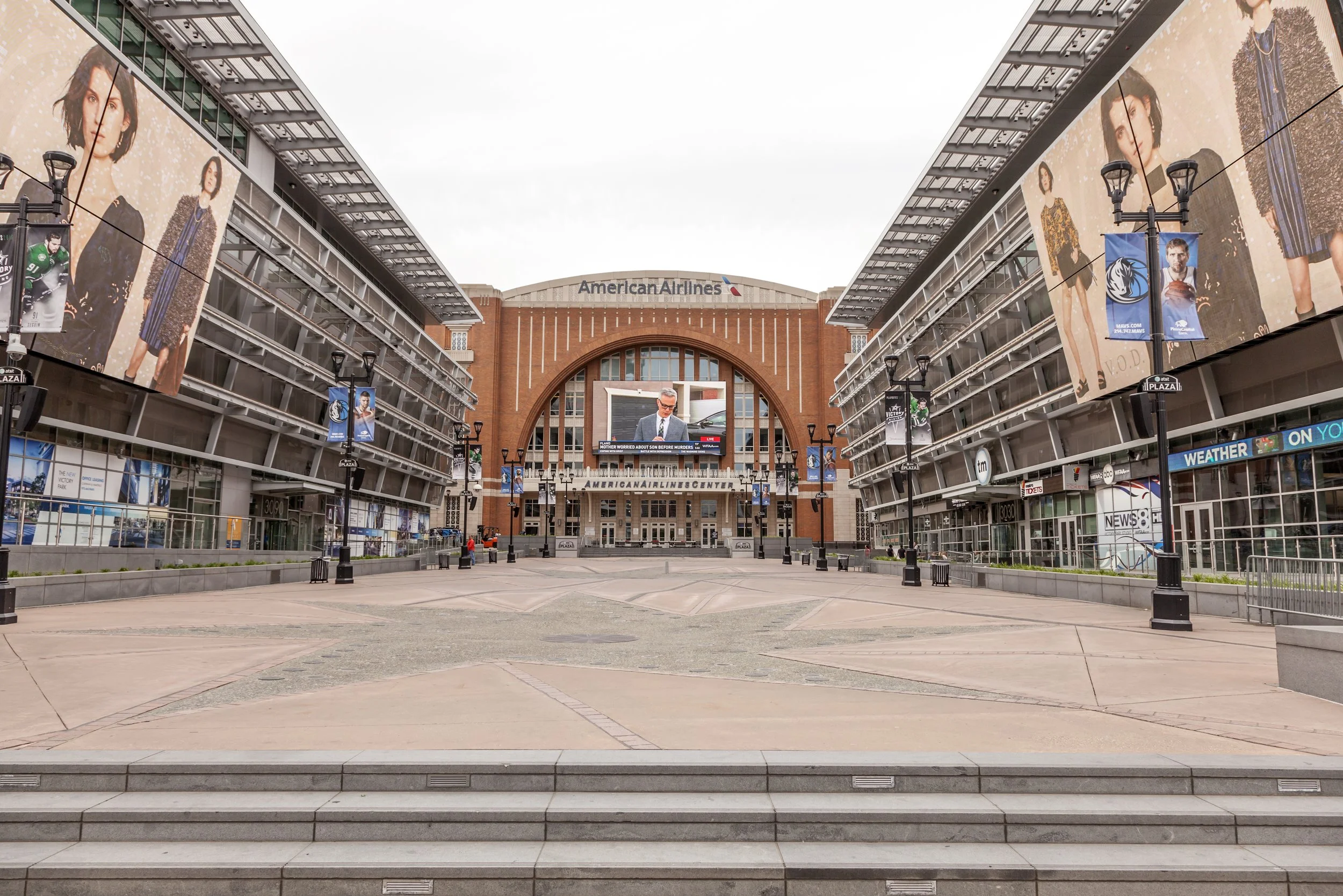 Empty outdoor plaza in front of American Airlines Center with large advertisements on both sides and a digital screen displaying a man in a suit.