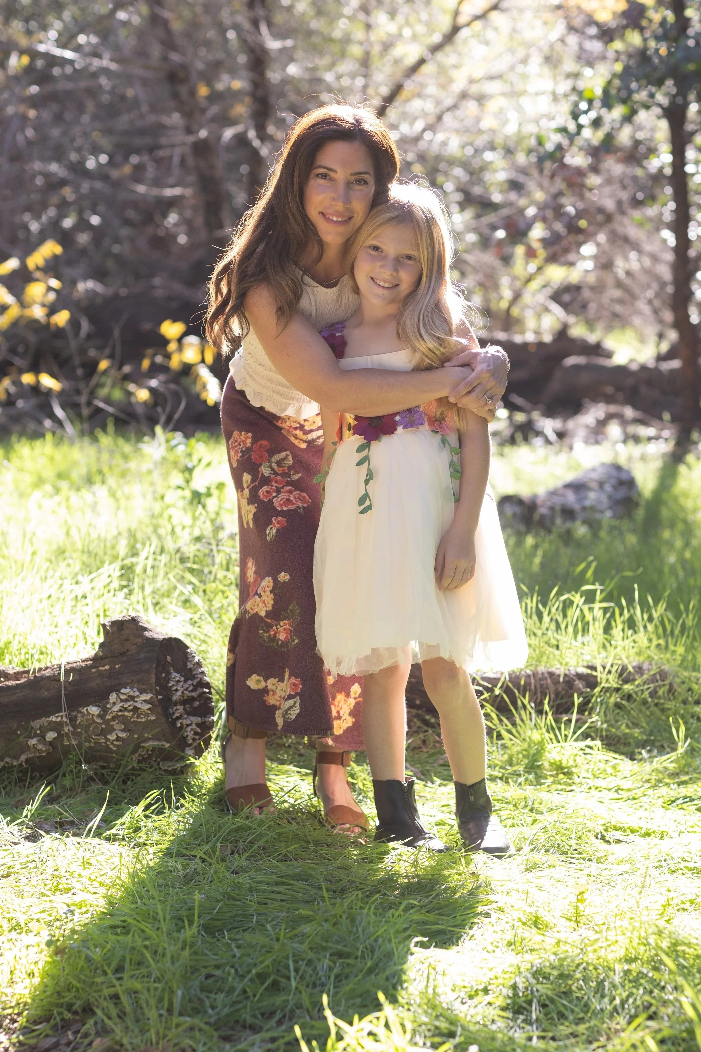 A woman and a young girl standing outdoors in a sunny, wooded area, hugging and smiling at the camera.