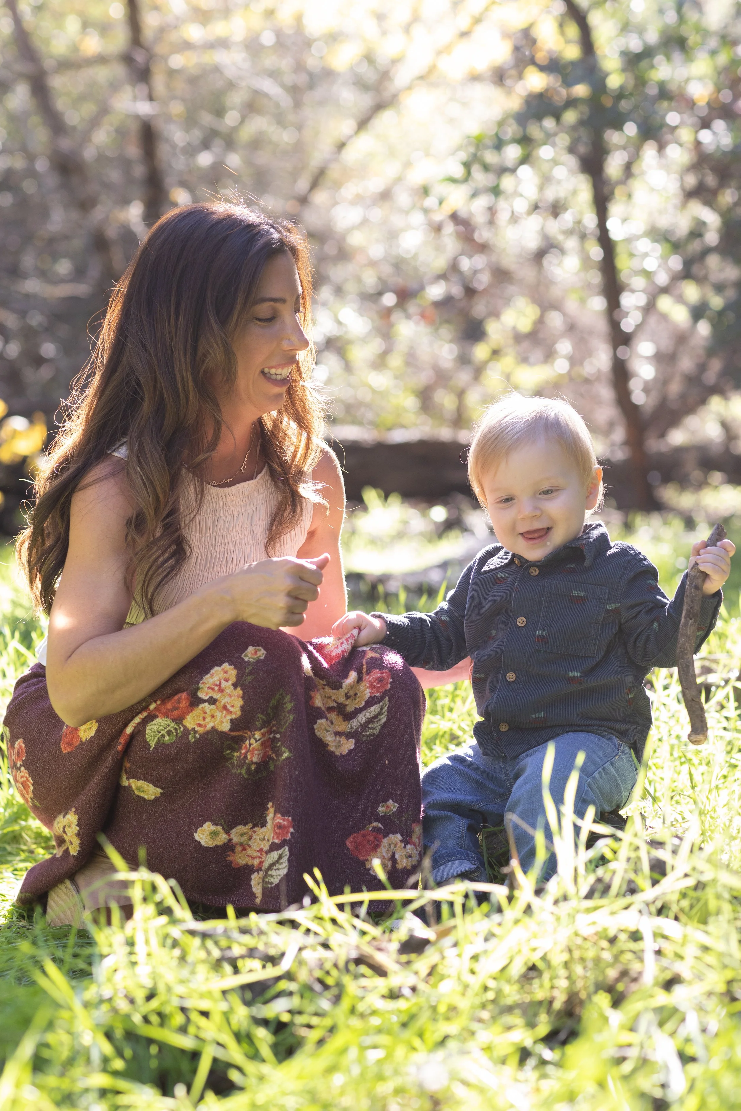 A woman with long brown hair and a young boy with light hair sitting outdoors in a grassy area, smiling and playing together with a stick, surrounded by trees with sunlight filtering through the branches.