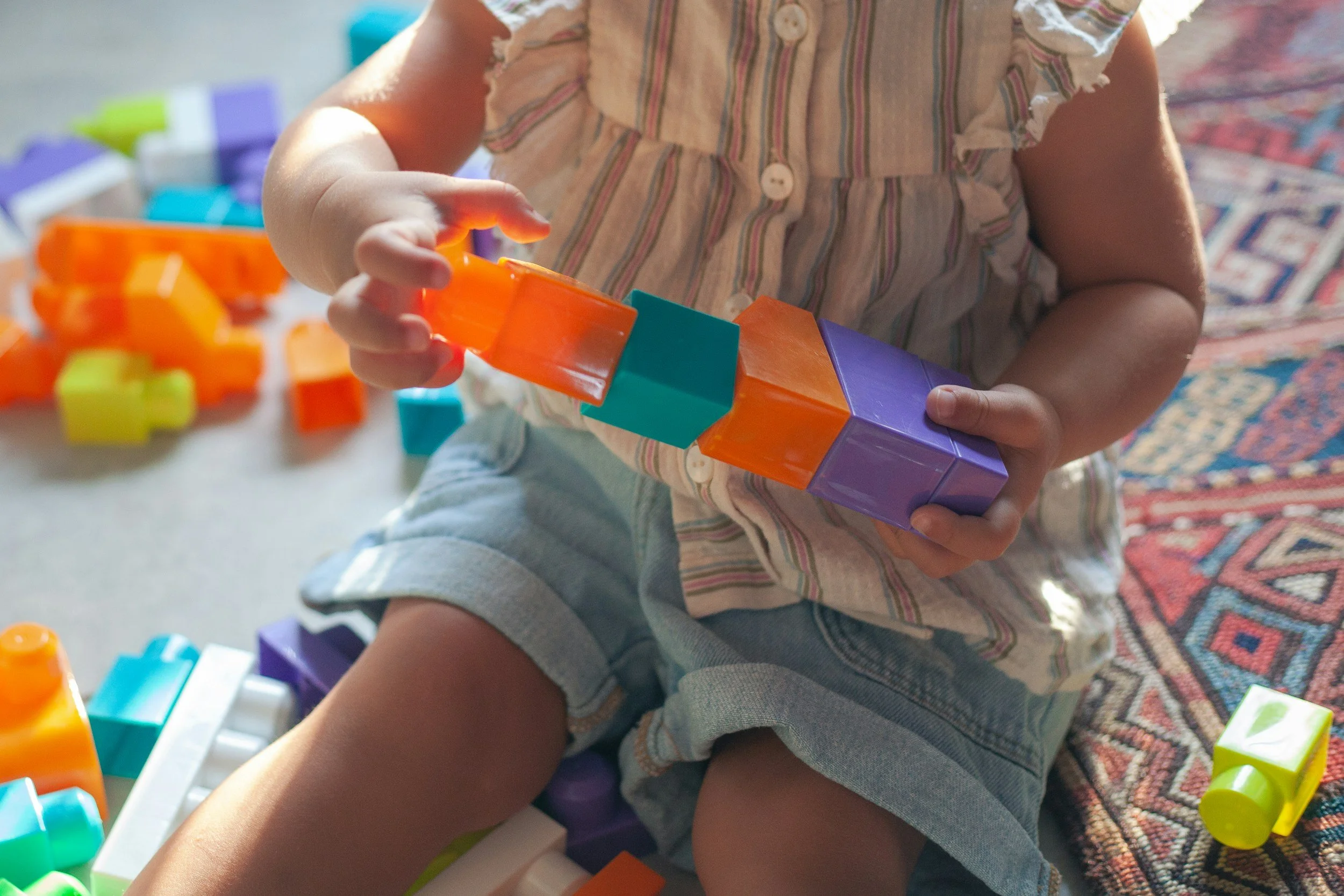 A young child plays with colorful building blocks, sitting on a patterned rug. The child's torso and hands are visible, wearing a striped shirt with distressed sleeves and denim shorts. The background shows scattered blocks.