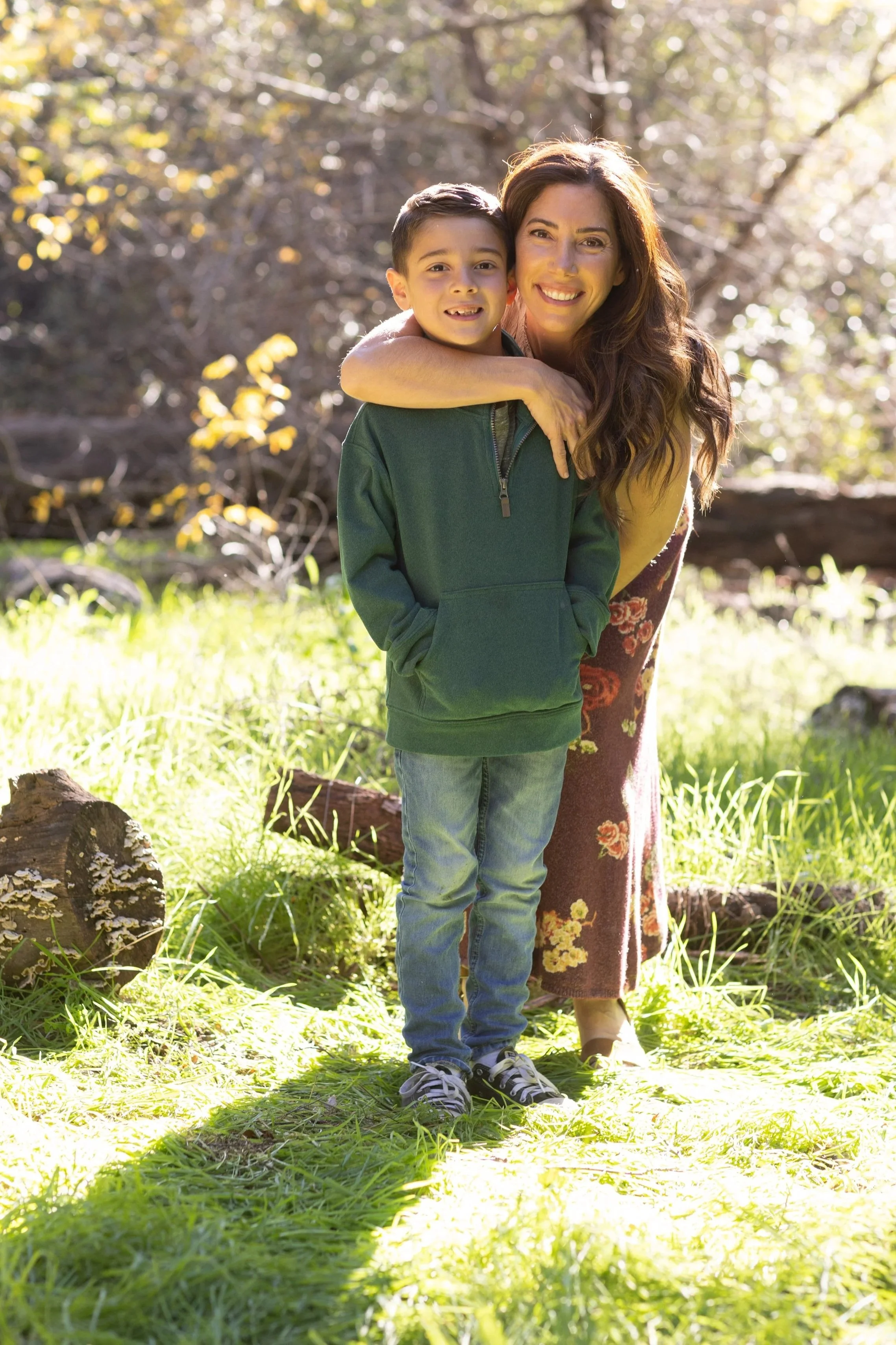 A woman with long brown hair and a young boy with short dark hair standing outdoors in a sunlit wooded area, smiling at the camera with trees and sunlight in the background.