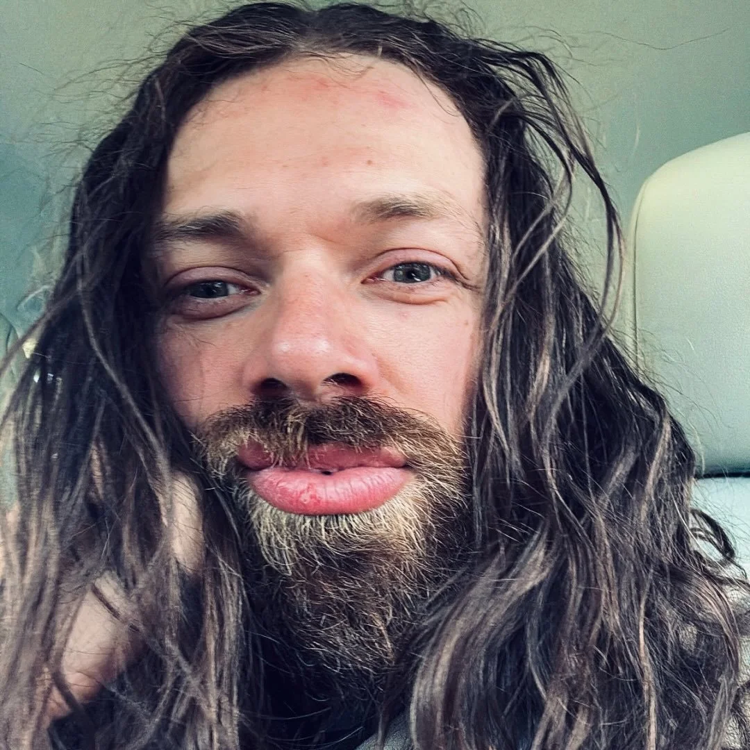 Close-up of a man with long wavy hair and a beard, looking at the camera with a slight smile.