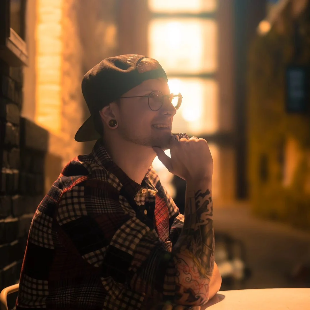 A young man with glasses, earrings, and tattoos sitting at a table, smiling while resting his chin on his hand in a warmly lit room with sunlight streaming through windows.