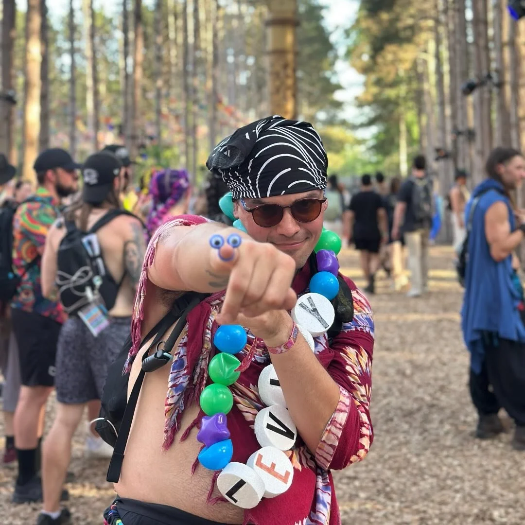 Man wearing sunglasses and a bandana, with colorful beads spelling 'LOVE' around his neck, pointing at the camera with a playful expression at a forest music festival.