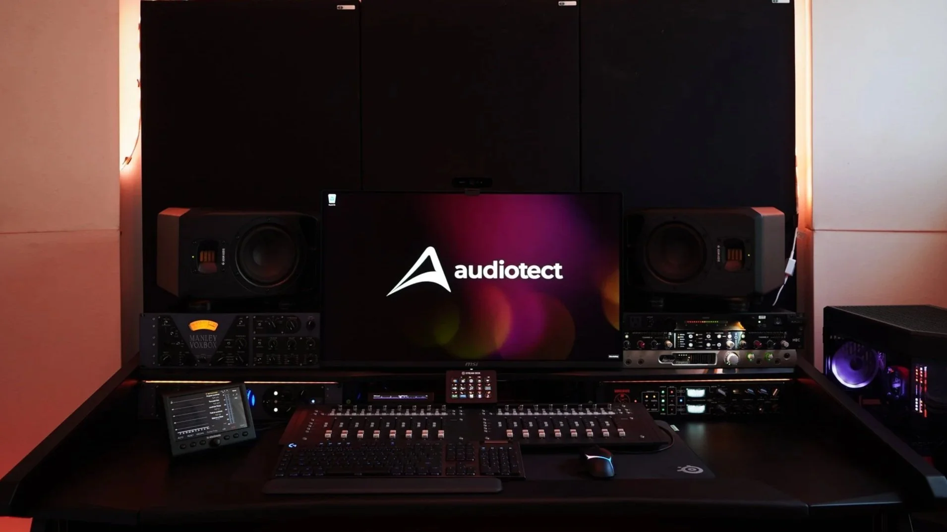 Photo of a music production studio setup with a computer monitor displaying the Audiotect logo, studio speakers, audio equipment, mixing console, and controls, with warm ambient lighting.
