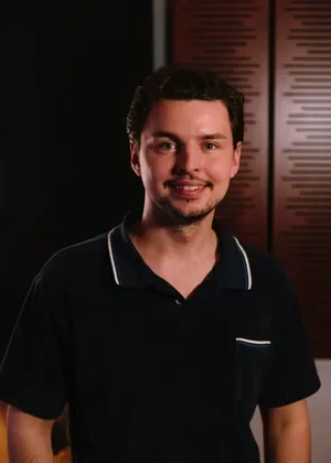 A young man with dark hair and a goatee smiling, wearing a black polo shirt with white trim, standing indoors in front of a dark background and a red menu board.