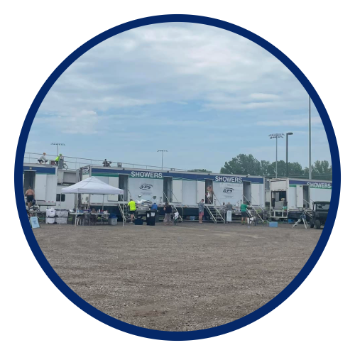 Image of a row of shower trailers with the word 'Showers' on each, set up outdoors on a gravel surface under a cloudy sky, with people and equipment nearby, possibly at a sporting or event venue.