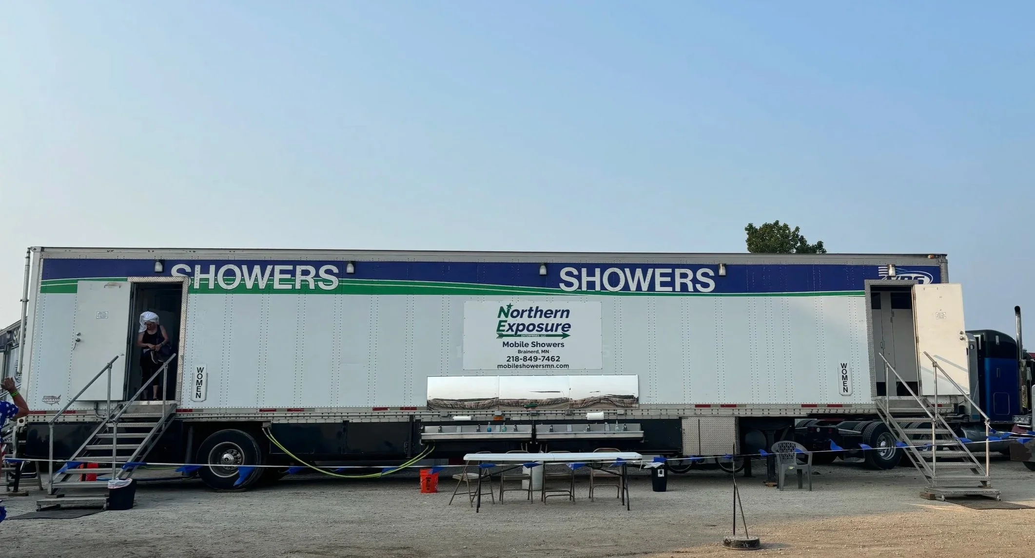 Mobile shower truck with signs for showers and Northern Exposure. A person with a white cap and black clothing standing at the door, stairs leading up to the vehicle, and some chairs and equipment around it.