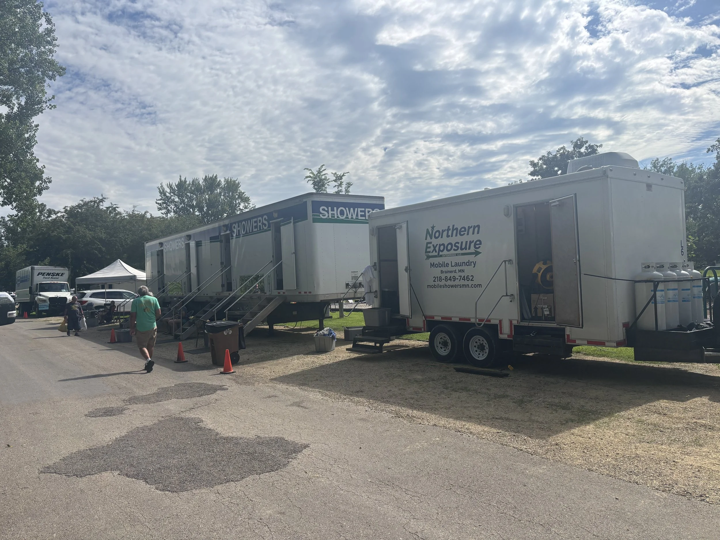 A mobile laundry trailer from Northern Exposure with a blue sign labeled 'Showers', set up outdoors with trees, tents, and people nearby under a partly cloudy sky.