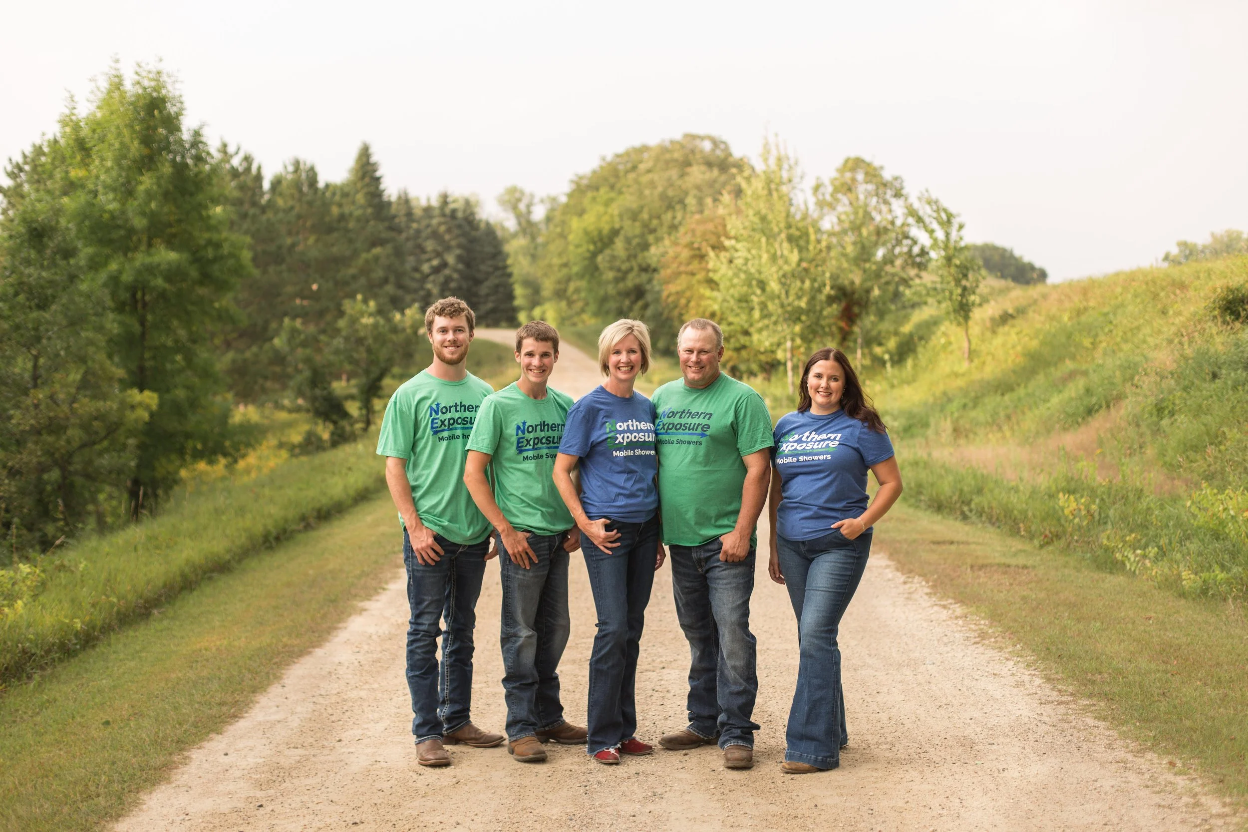 Group of five people standing on a dirt path surrounded by green trees, wearing matching T-shirts with 'Northern Exposure Mobile Showers' printed on them.