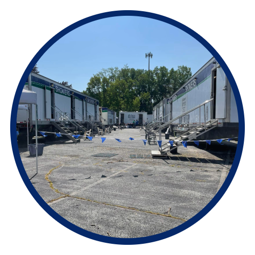 Outdoor trailer parking lot with trailers on both sides, set up as a shower station area with portable showers, stairs, and blue bunting, under a clear blue sky.