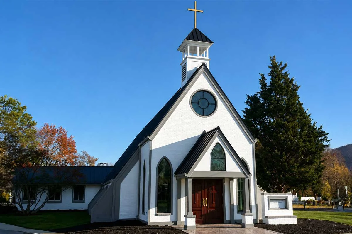 White church with black roof, prominent cross on steeple, arched windows, and surrounded by trees and a well-kept lawn.